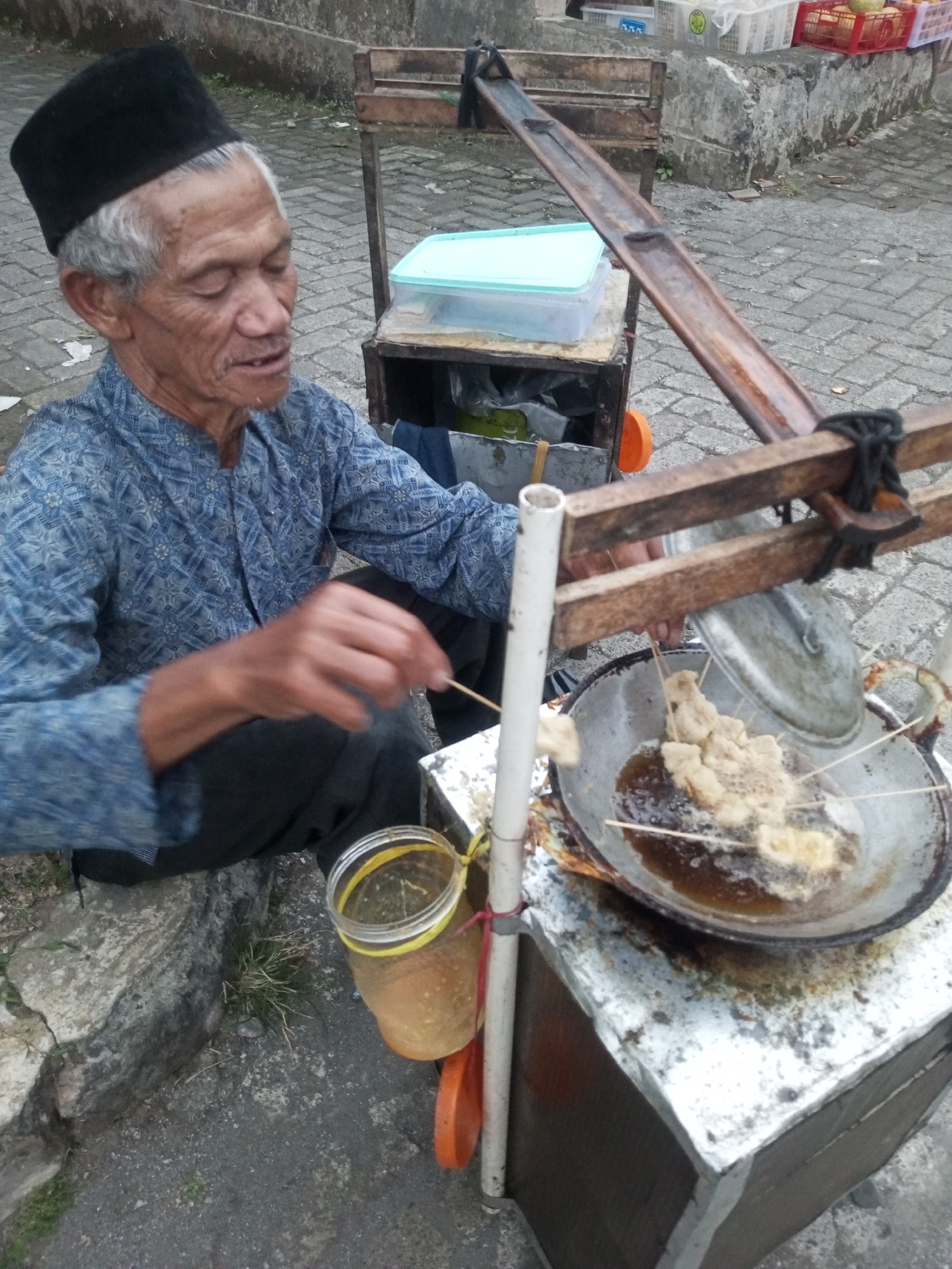 An elderly Indonesian man with graying hair, wearing a black peci cap and a blue patterned long-sleeved shirt, is seen from a high angle, preparing food at a street cart. He is seated with his legs crossed, and his right hand is raised, holding a skewer of battered and fried food that is immersed in a wok filled with hot oil. The wok is placed on a metal cart, which also holds a clear plastic container with a light blue lid and a glass jar filled with a yellowish liquid, likely oil or sauce. The street cart appears to be made of metal and wood, with a bamboo-like structure extending upwards, possibly for hanging items or supporting the cooking apparatus. The ground is paved with cobblestones, and a rough stone wall is visible in the background. In the distance, several plastic crates containing produce can be seen, suggesting a market or food stall area. The overall scene depicts a typical street food vendor in Indonesia, likely in the city of Andongsili, given the context of the prompt.