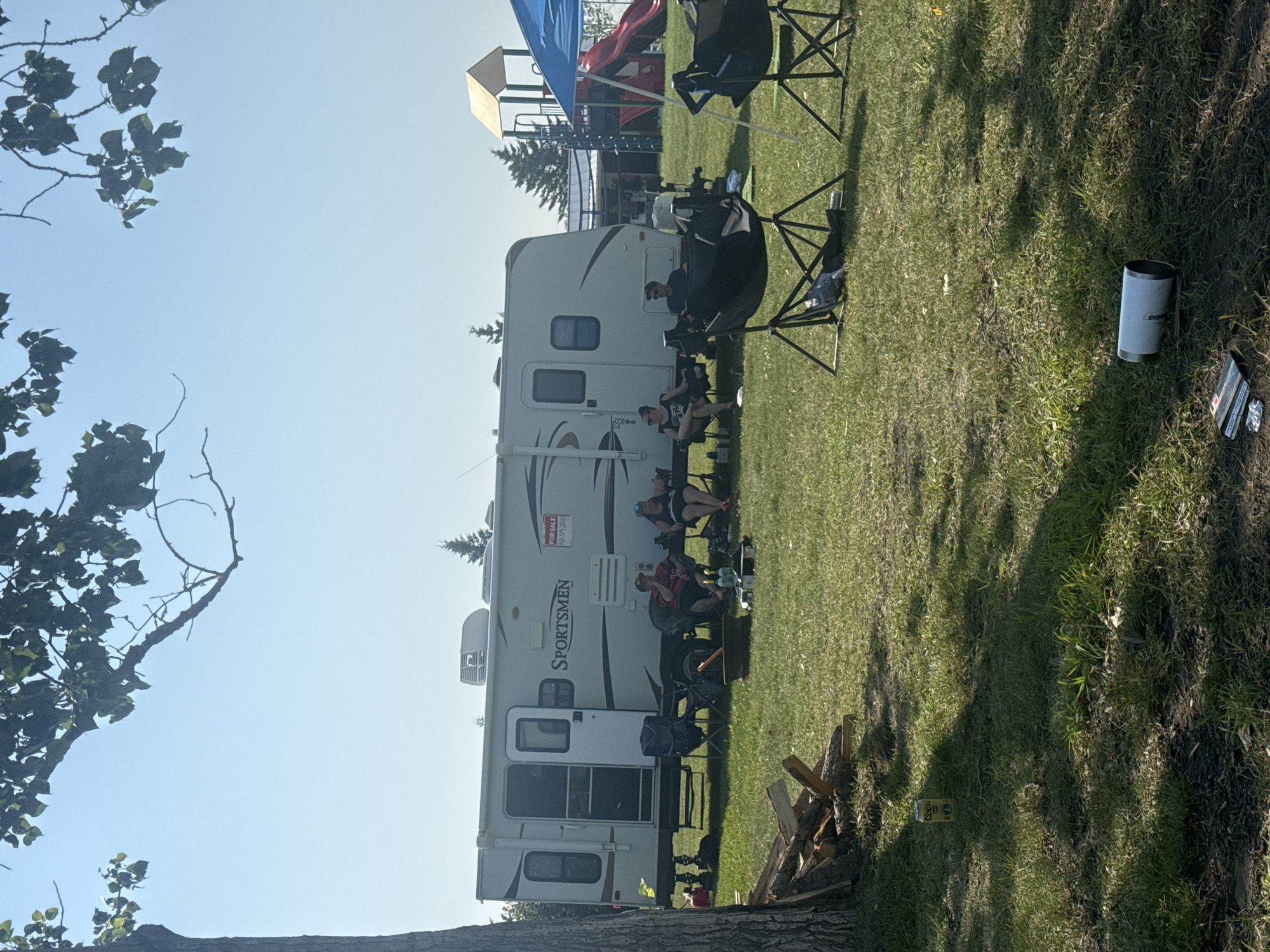 The image depicts an outdoor scene at a campsite in Lyalta, Alberta, Canada. A large white recreational vehicle (RV), labeled "SPORTSMEN," is parked in the center, with its door open. A "FOR SALE" sign is visible on the side of the RV. Several people are seated in lawn chairs around the RV. To the left of the RV, a pile of firewood is stacked near a tree. In the foreground, the grass is green and somewhat dry in patches. Several empty lawn chairs are scattered around. A blue canopy is visible on the right side of the frame, with a red slide and playground structure in the background. A single white can, identified as "Coors Light," sits on the grass to the left. Further to the right, a white tumbler and some crumpled foil are on the ground. The sky is clear and blue, with branches of a tree hanging into the frame from the top. The overall impression is of a casual, relaxed outdoor gathering at a campsite.