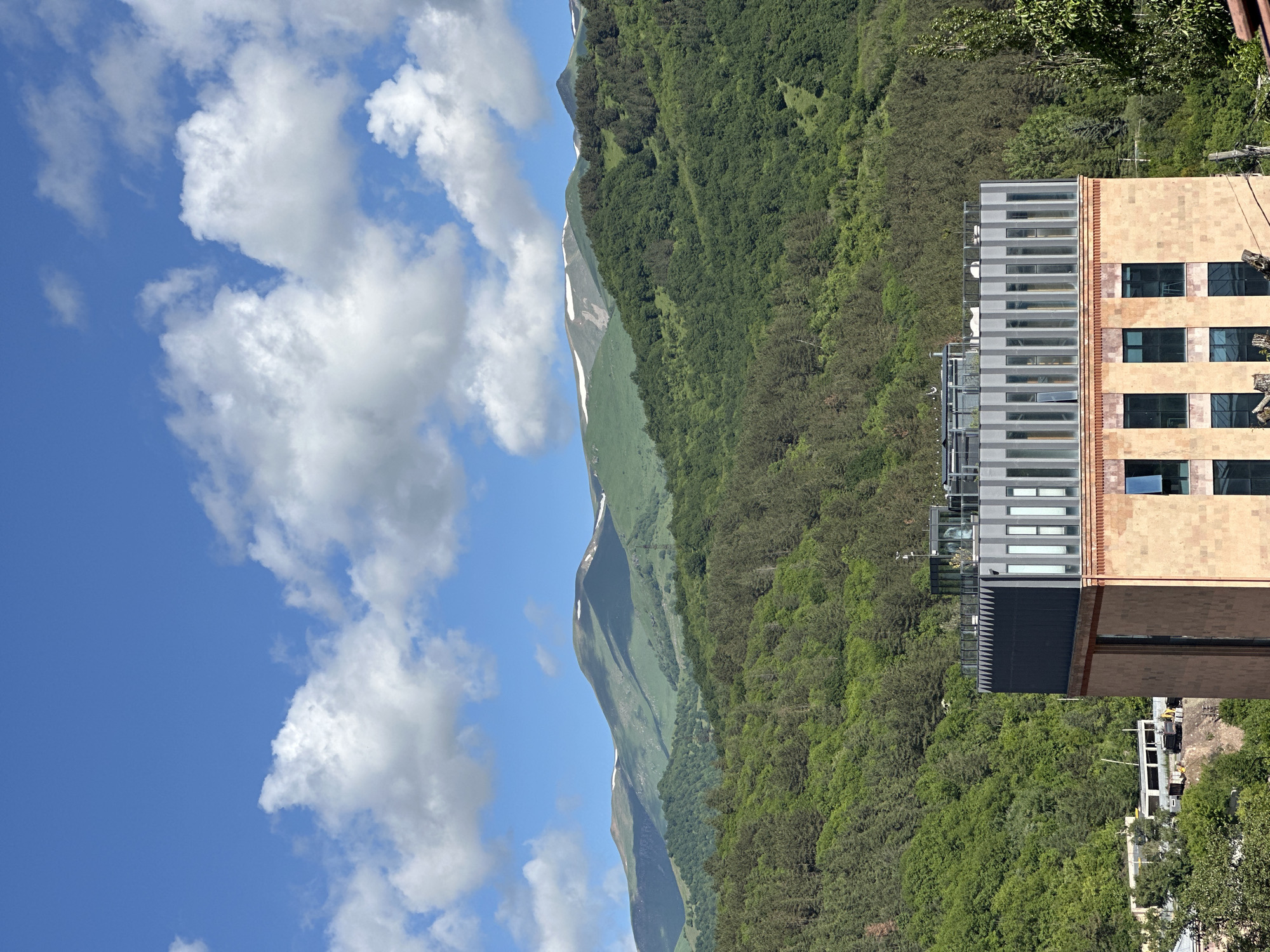 The image captures a scenic view in Dilijan, Armenia, dominated by lush green mountains and a bright blue sky dotted with white clouds. In the foreground, a modern building with a terracotta-colored facade and a metallic upper story is partially visible. The building features large windows, some of which are open, revealing glimpses of the interior. The metallic upper section is adorned with vertical glass panels and has an open-air rooftop area with some white seating.
Beyond the building, dense forests cover the slopes of the mountains, with a few patches of snow visible on the higher peaks in the background. The mountains are layered, creating a sense of depth and vastness. The overall scene suggests a peaceful and picturesque environment. In the lower left of the image, a construction site with heavy machinery and some smaller buildings is visible, hinting at ongoing development in the area.