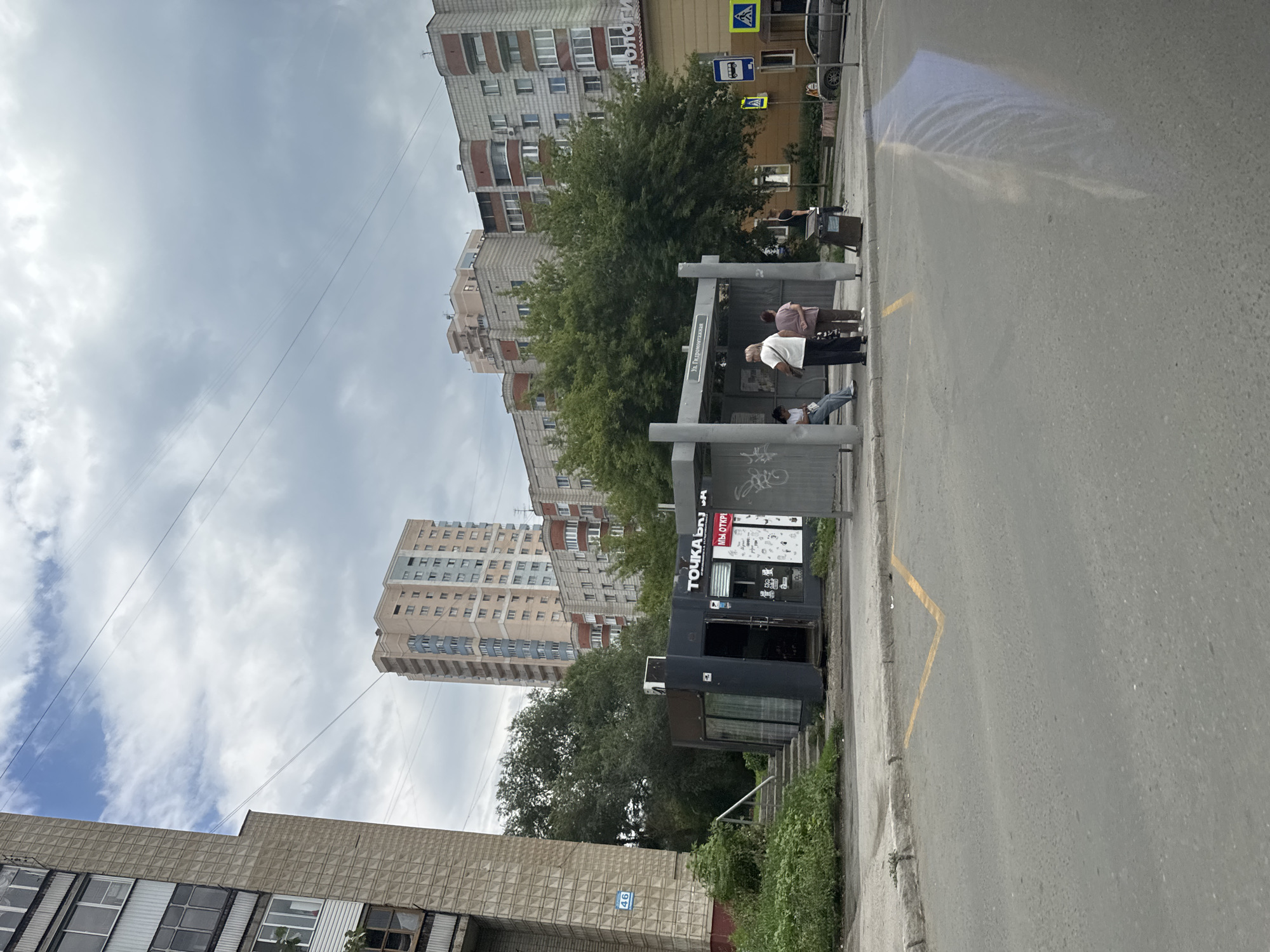 In this medium-shot, outdoor photograph, a bus stop shelters are present on a cloudy day. On the left, a utilitarian, gray kiosk with glass windows and a dark-colored entrance stands on a concrete platform, with a set of concrete stairs leading up to it. Adjacent to the kiosk, a tall, beige apartment building with numerous windows dominates the background. In the center of the frame, a bus stop shelter with metal poles and a gray roof provides cover for several individuals. A sign above the shelter reads "Ул. Гидромонтажная." Several people are standing or sitting under the shelter, awaiting transportation. To the right of the bus stop, a yellow and orange building with windows and a sign that reads "ТОПОЛОГИ" is visible. A street with yellow lane markings stretches across the foreground. A car is parked on the right side of the road. The environment appears urban, with signs of wear and tear on the infrastructure.