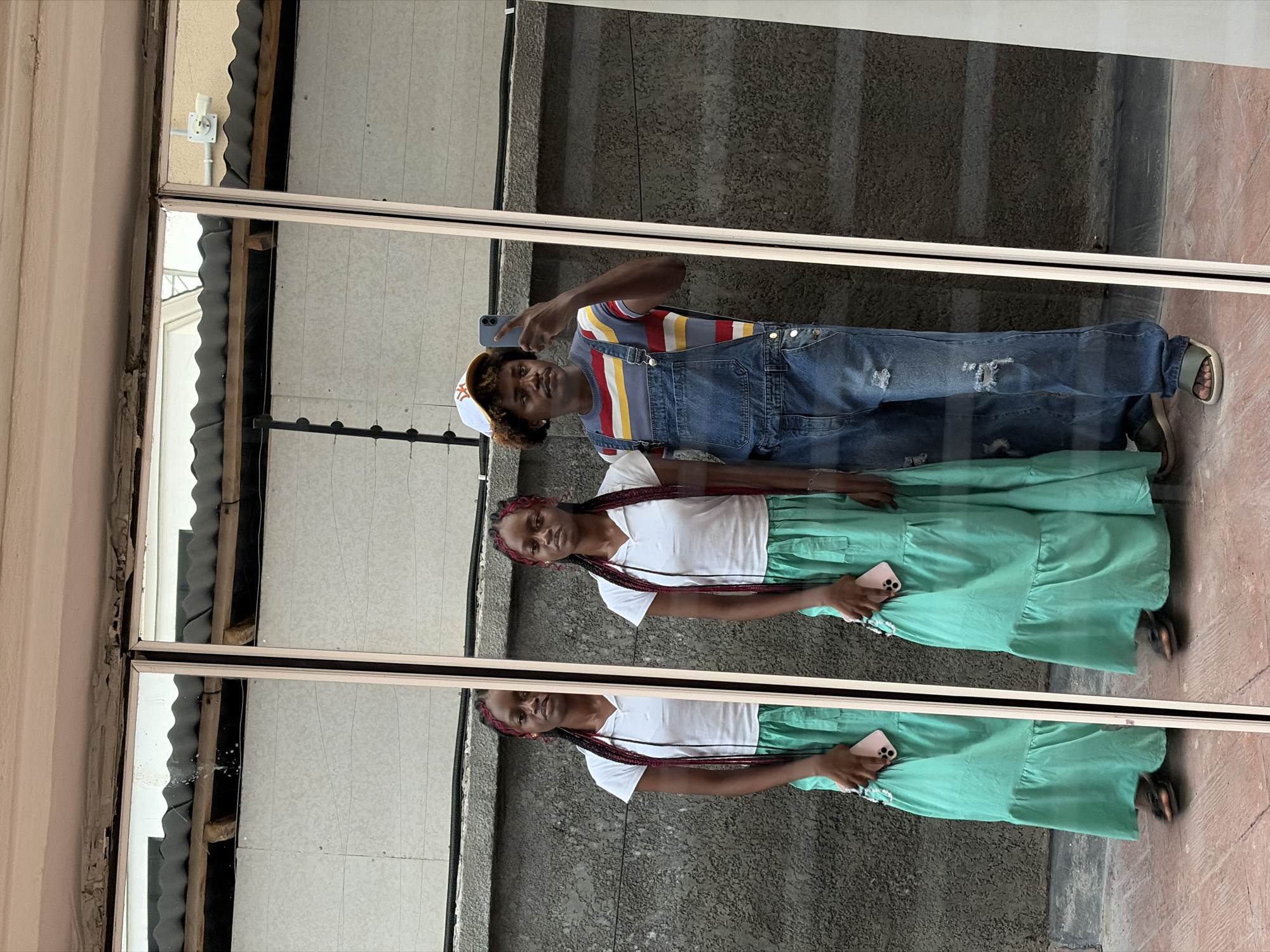 The image captures two individuals, a male and a female, in what appears to be a mirror selfie or a photo taken through a highly reflective window in Igboefon, Nigeria. The image is rotated 90 degrees clockwise.
The male subject stands behind and slightly to the right of the female. He is holding a light blue smartphone up, seemingly taking the photo. He wears a blue and white striped t-shirt with red and yellow accents, blue denim overalls with distressed patches on the knees, and light-colored sandals. A white baseball cap with a prominent "NY" (New York Yankees) logo covers his curly hair. He looks directly at the camera.
The female subject is in the foreground, holding a light pink smartphone in her left hand. She is dressed in a plain white t-shirt and a long, flowing light green tiered skirt, paired with dark sandals. Her dark hair is styled in long braids featuring distinct red streaks. She also looks towards the camera.
The background behind the subjects is a dark, textured wall, possibly concrete or stucco, with a lighter, tiled floor below. The reflections in the mirror/window reveal parts of the external or adjacent environment, including corrugated metal roofing, a white electrical fixture, and structural elements of a building. The bright lighting suggests it is daytime.