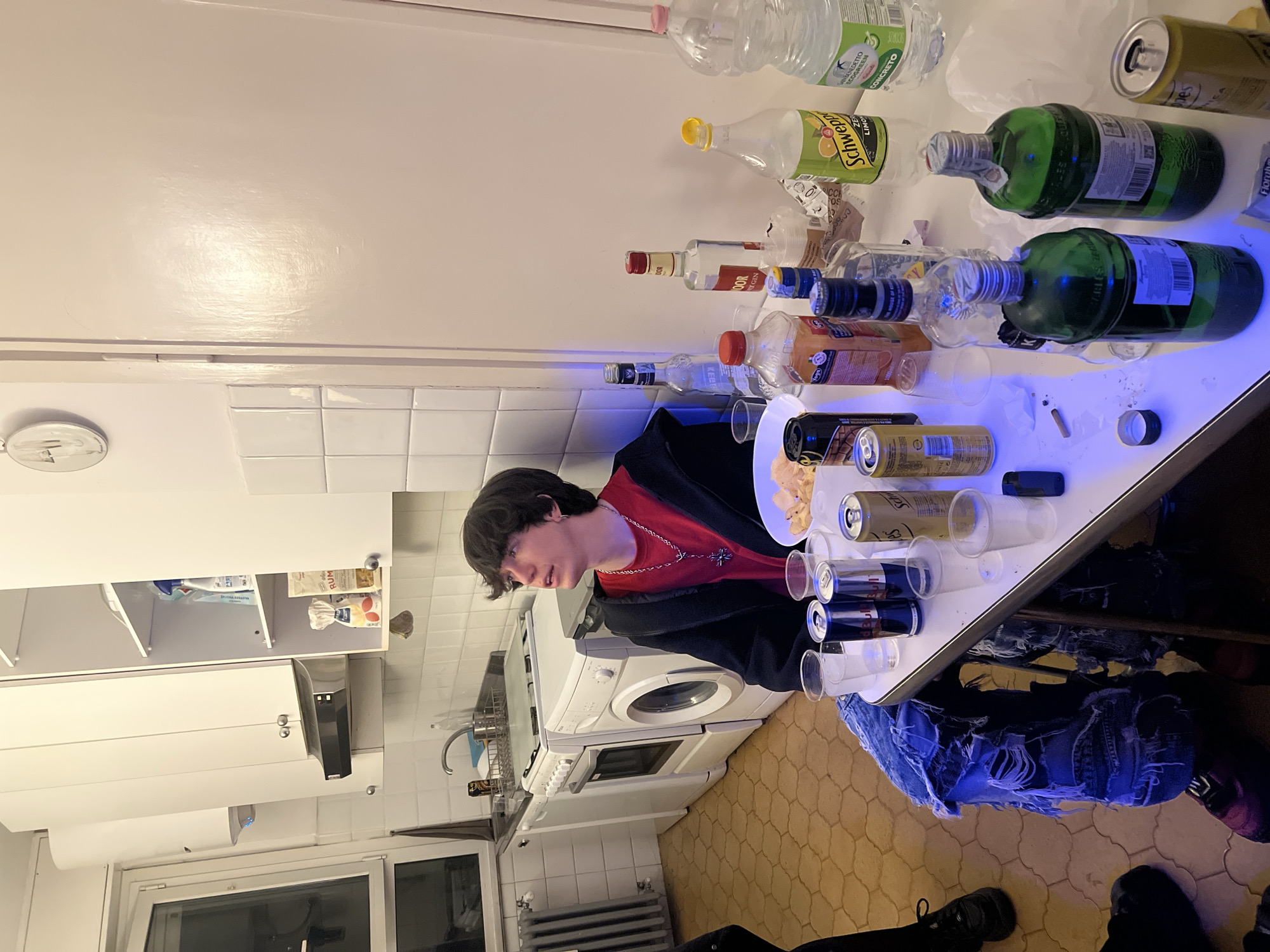 A young man with dark hair stands in a kitchen, facing a table covered with empty bottles and cans. The bottles include water, Schweppes, and several liquor bottles. There are also glasses and a plate of chips on the table. The kitchen features white cabinets and tiled walls, and appliances like a washing machine and stove. The floor is tiled in a geometric pattern. The scene appears to be indoors. There is no location in Milan, Italy identified.