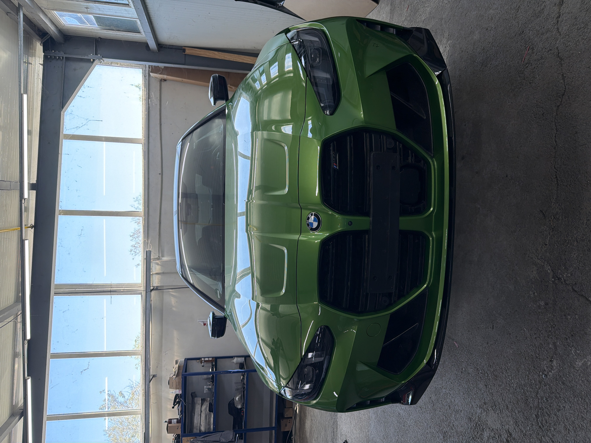 A frontal view shows a green BMW automobile in a garage or automotive shop setting. The car faces directly towards the camera, showcasing its grille, headlights, and the BMW emblem. Overhead, a large window complex is visible, possibly allowing natural light to enter the garage. Behind the car, a shelving unit is present, containing various items; the background shows details of the garage structure, including beams and walls. The concrete floor is unadorned. There is no information to indicate a location in Burgas, Bulgaria. The scene appears unoccupied, focusing solely on the vehicle and its immediate environment.