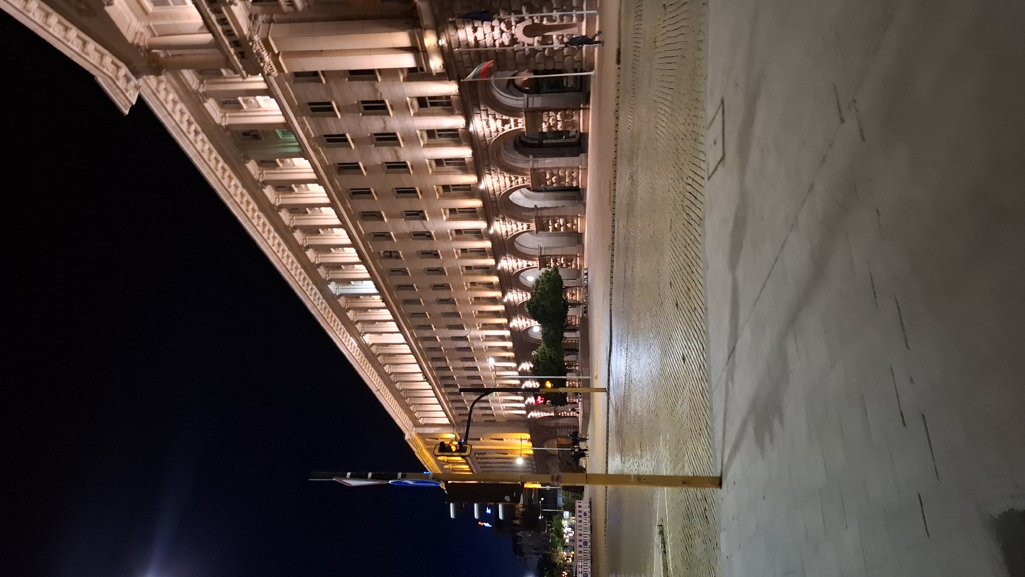 The image captures a nighttime view of the Largo complex in Sofia, Bulgaria, specifically focusing on the Presidency building. The building, a grand structure with classical architectural elements, is illuminated by warm exterior lights, highlighting its numerous windows and arched ground floor. The street in front of the building is paved with cobblestones, reflecting the ambient light. A traffic light, displaying yellow, stands prominently on the left side of the image, with a yellow pole in the foreground. Several trees are visible along the building's facade, their branches silhouetted against the illuminated stone. A Bulgarian flag flutters from a flagpole in front of the building. A lone figure, appearing as a pedestrian, walks on the sidewalk near the flag. The sky is dark, indicating the late hour. The overall scene conveys a sense of urban tranquility at night.