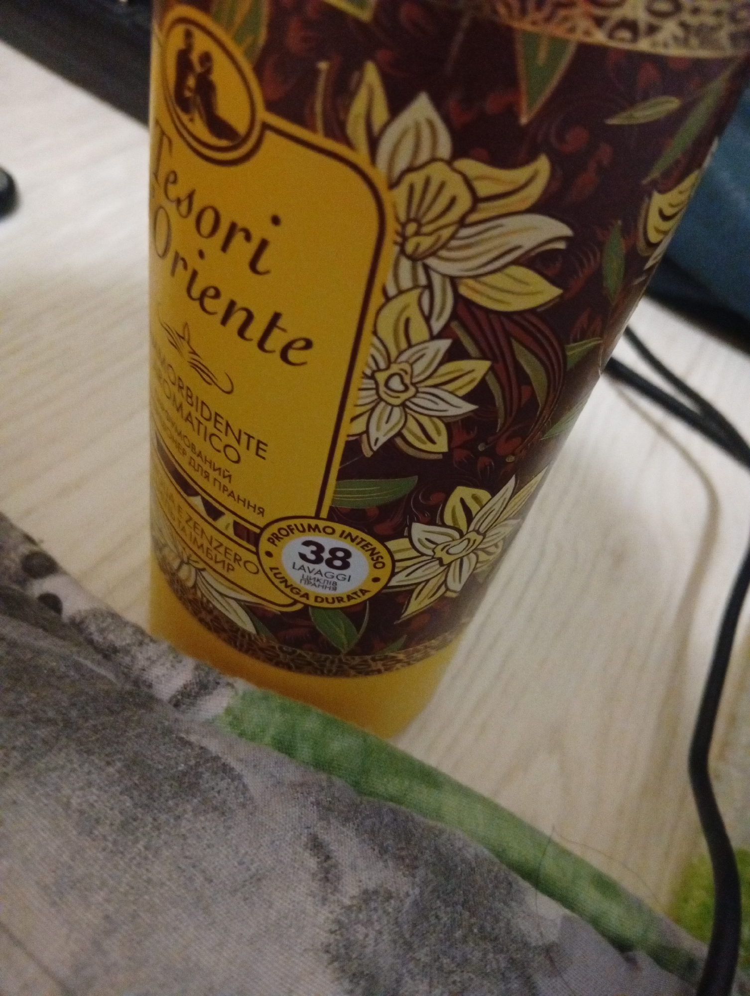 The image shows a partially visible bottle of "Tesori d'Oriente" fabric softener, specifically the "Aromatic" variant with "Zenzero e Seta" (Ginger and Silk) scent. The packaging is predominantly yellow with floral designs. A circular label indicates "Profumo Intenso 38 Lavaggi Lunga Durata" (Intense Perfume 38 Washes Long Lasting).
The bottle is placed on a light-colored surface, likely a table or countertop. A gray and green patterned fabric is visible in the lower left corner. A black cable is present on the right side of the frame. The image does not contain sufficient detail to determine if the image was taken in Poltava, Ukraine.
