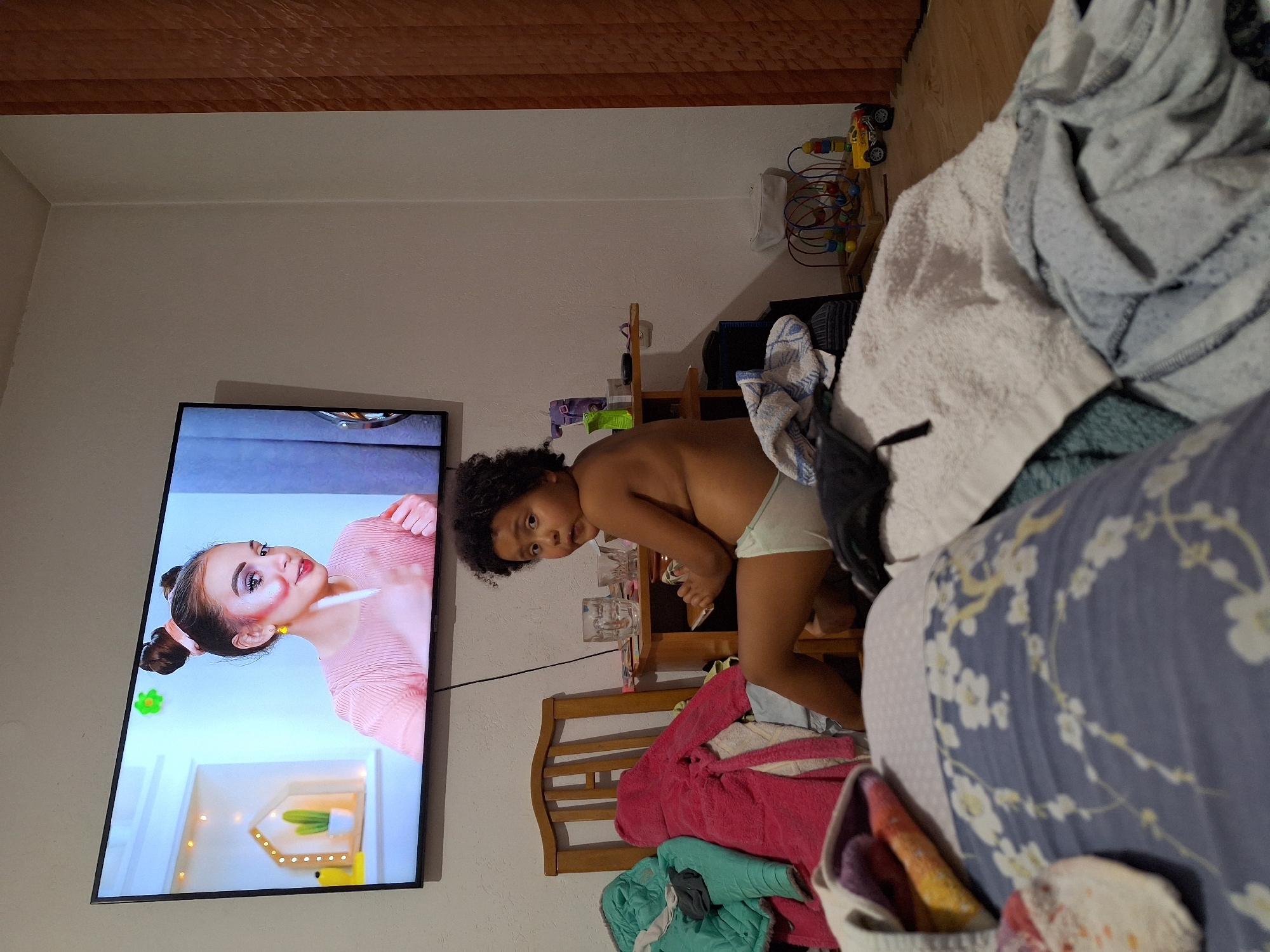 The image captures an indoor scene, likely a living room, where a young child is seated on the floor in front of a television. The television screen displays a young woman applying makeup. The child, with curly dark hair, is wearing a light-colored diaper and is looking towards the camera with a surprised expression. The floor is covered with a large pile of laundry, including towels and blankets, one of which has a floral pattern. A wooden chair and a small table are visible behind the child. The table holds two glasses and some small items. In the background, a toy vehicle and a bead maze toy are on the floor. The overall impression is one of a casual, lived-in domestic environment.