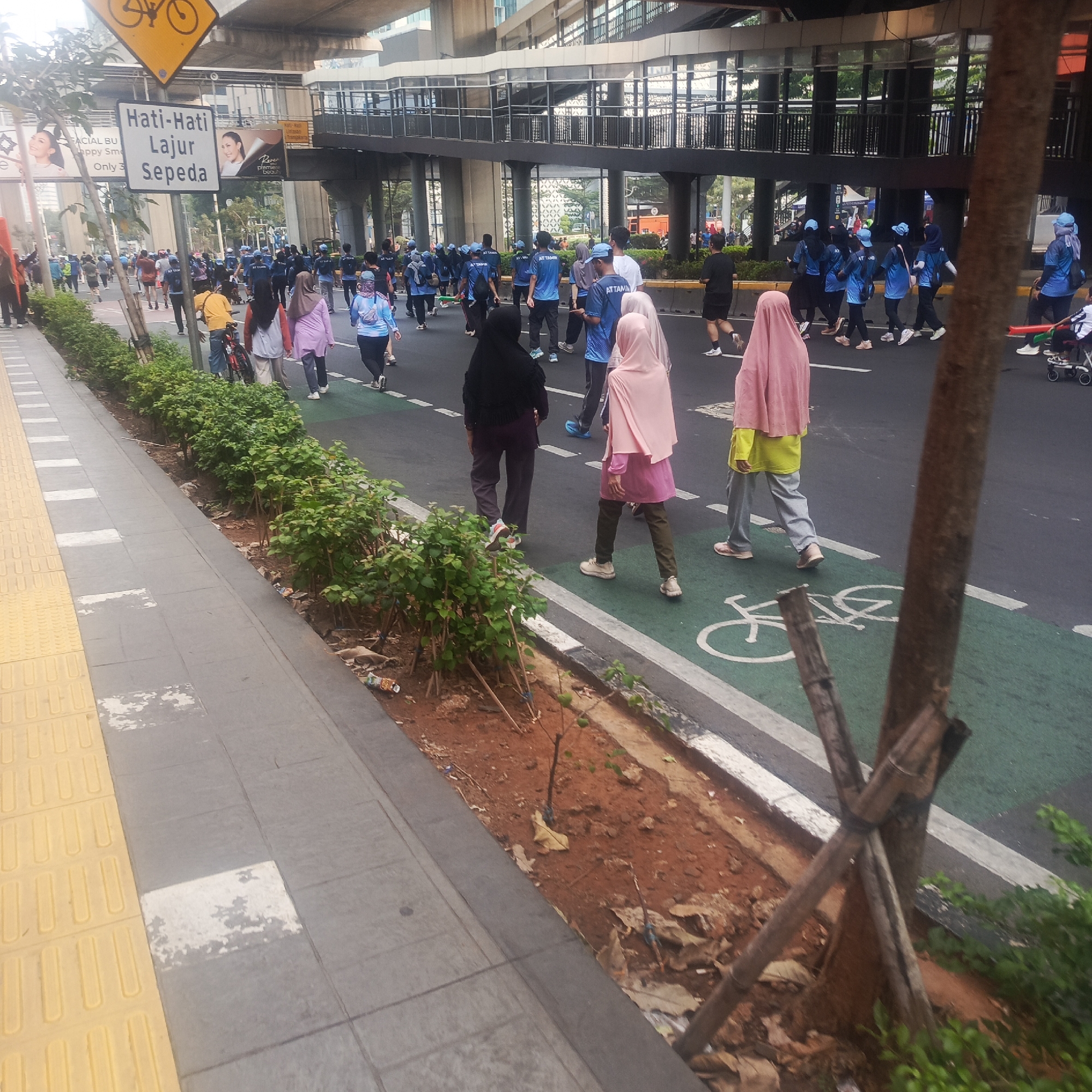 In South Jakarta, Indonesia, a group of people are participating in an outdoor event, likely a fun run or walk, on a wide street with designated lanes for cycling and pedestrian traffic. The scene is urban, with a modern pedestrian overpass and elevated train tracks visible in the background, suggesting a busy thoroughfare.
The foreground shows a pedestrian walkway with textured yellow tactile paving for the visually impaired, alongside a gray paved area with white markings. Adjacent to the walkway is a strip of reddish-brown soil with young plants and some scattered litter. The street itself is paved with asphalt and features a green lane marked with a white bicycle symbol, indicating it's a dedicated bike path.
The participants are a mix of individuals, many of whom are wearing matching blue t-shirts with the text "ATTAMIN" printed on them, suggesting they are part of a team or organization. Some are wearing headscarves and long-sleeved shirts, indicating adherence to modest dress. Their attire is casual, suitable for physical activity, with many wearing athletic shoes.
The people are generally walking or jogging, with some interacting with each other. One individual is seen pushing a stroller. The overall atmosphere appears to be one of community and shared activity. The time of day seems to be morning or afternoon, with the lighting suggesting daylight. The environment is clean overall, with the exception of some minor litter along the roadside. A sign with Indonesian text, "Hati-Hati Lajur Sepeda," which translates to "Beware of Bicycle Lane," is prominently displayed on a pole in the foreground, along with a yellow sign depicting a bicycle symbol.