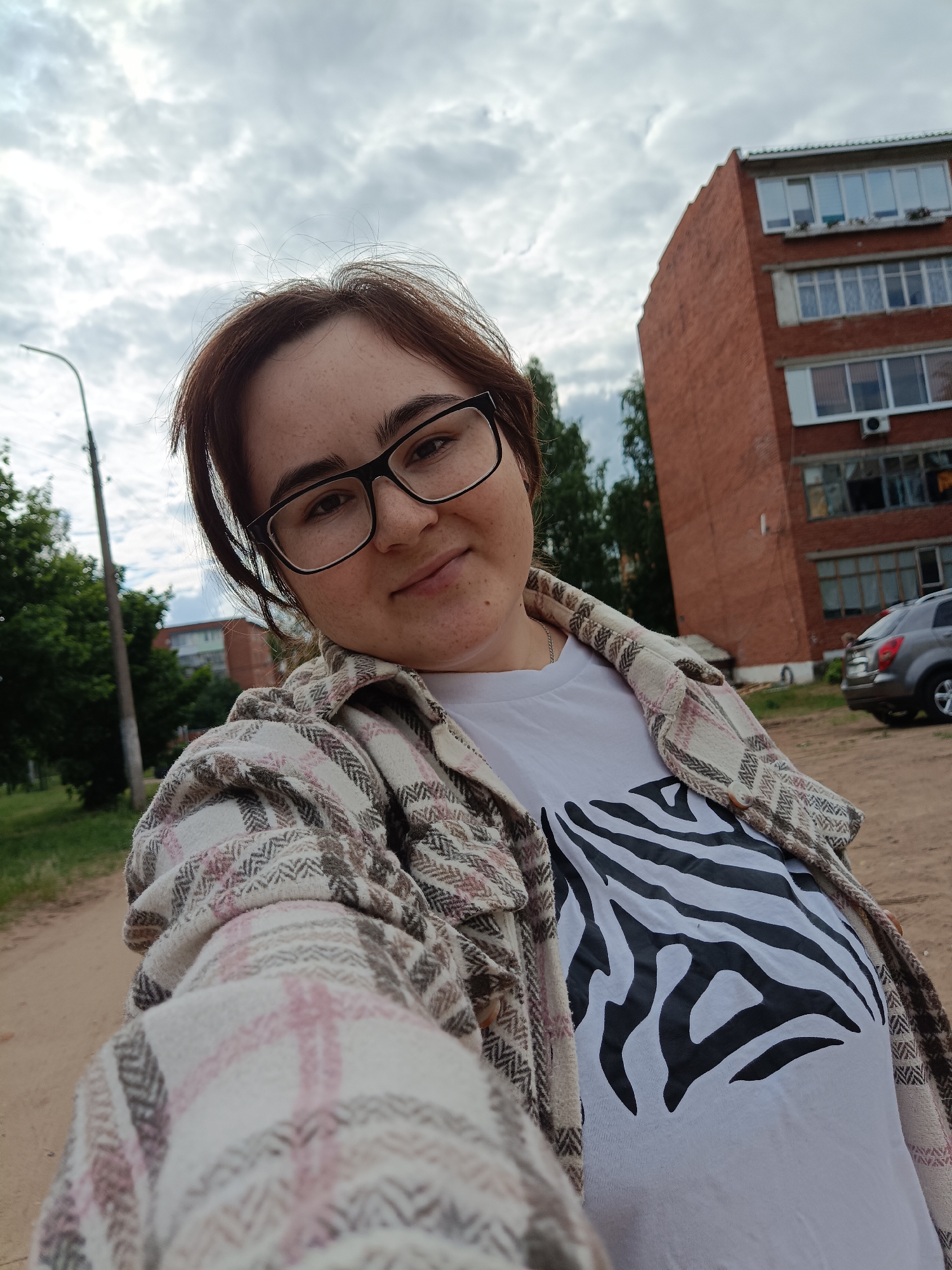 A selfie taken outdoors in Noviy, Russia, features a person with dark hair, black-framed glasses, and freckles, looking directly at the camera with a gentle smile. She is wearing a white t-shirt with a bold black zebra stripe pattern, layered under an open, long-sleeved plaid shirt in shades of white, pink, and brown.
The setting is a residential area on what appears to be an overcast day, indicated by the cloudy sky. In the background, a tall street light pole and lush green trees are visible to the left. To the right, a multi-story brick apartment building with numerous windows stands prominently. A grey SUV is parked on the unpaved ground in the mid-ground. The ground itself is a mix of dirt and gravel. The overall scene suggests a casual moment captured in an urban or suburban environment. There is no visible text in the image.