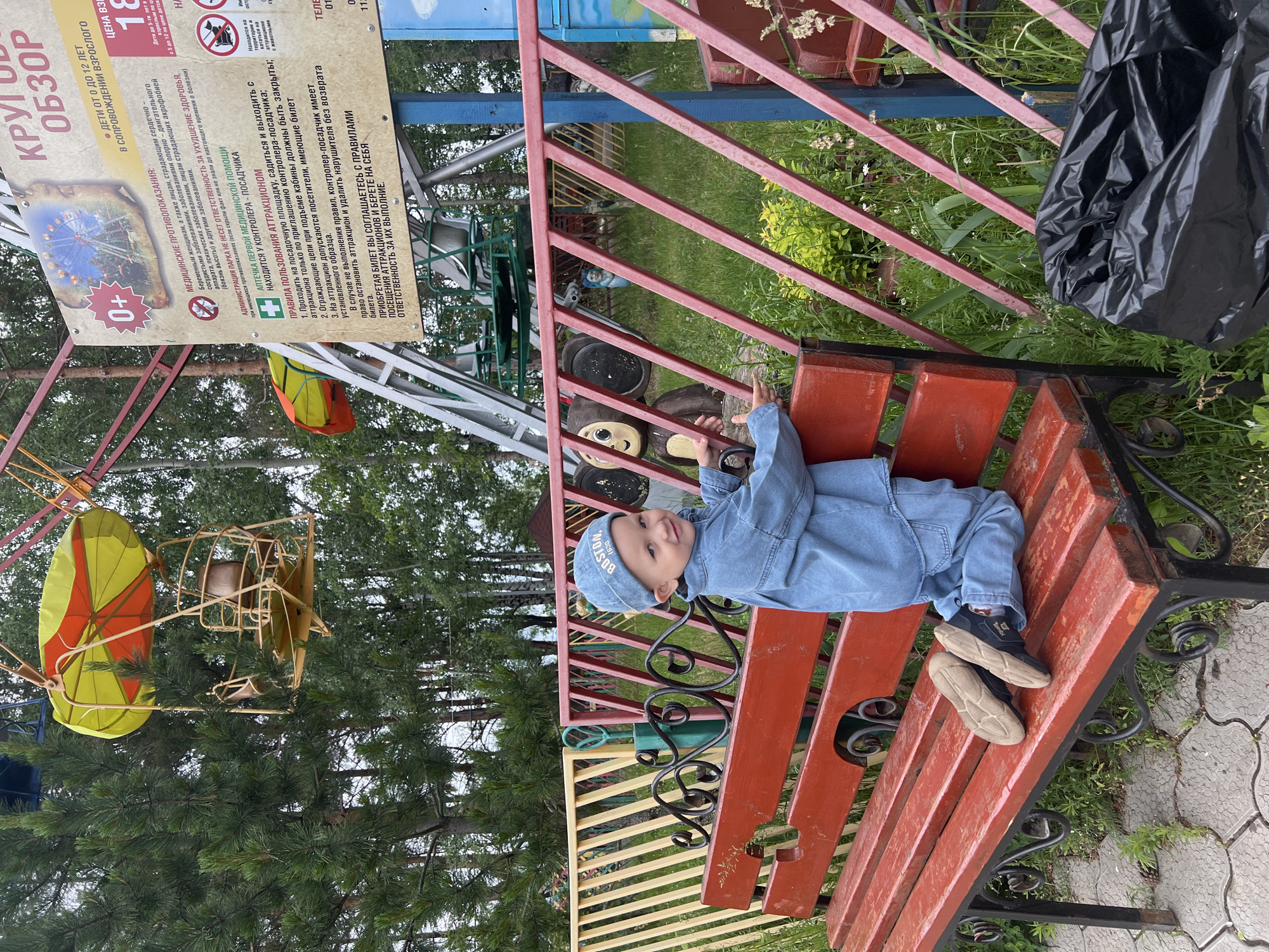 The image captures a young child posing on a park bench in an amusement park located in the city of Chaikovsky, Russia.
In the foreground, a young child, appearing to be a boy, kneels on a weathered red wooden slatted park bench with black wrought iron supports. He is dressed in a light blue denim jacket and matching trousers, along with a light blue cap embroidered with "BOSTON 1630" in white text. His dark blue sneakers with light brown soles are also resting on the bench. The child is looking directly at the viewer with a slight smile, his left hand gripping the top rail of a red metal fence positioned directly behind the bench.
Behind the red fence, which features diagonal and horizontal bars, a yellow and green striped fence is partially visible. Further back, on the right side of the frame, a large white sign with Russian text provides information about an amusement ride. The sign indicates a "0+" age rating, medical contraindications, park administration details, first aid information, and attraction rules. It features a graphic depicting a Ferris wheel and mentions "КРУГ ОБЗОР" (Observation Wheel) and "ДЕТИ ОТ 0 ДО 12 ЛЕТ В СОПРОВОЖДЕНИИ ВЗРОСЛОГО" (Children from 0 to 12 years accompanied by an adult). To the left of the sign, a brown monkey-like figurine is partially visible behind the red fence.
In the midground and background, an observation wheel or Ferris wheel is prominent. One of its yellow and orange umbrella-topped gondolas is visible in the upper left, suspended amongst tall pine and deciduous trees. Parts of the ride's metallic support structure are also seen. To the right of the main sign, parts of another green-colored amusement ride structure are discernible. The environment is lush with green grass, various plants, and wildflowers growing around the park features. The ground directly beneath the bench is paved with light-colored interlocking concrete pavers. In the lower right corner, a black plastic garbage bag is visible on the ground. The lighting suggests an overcast day.