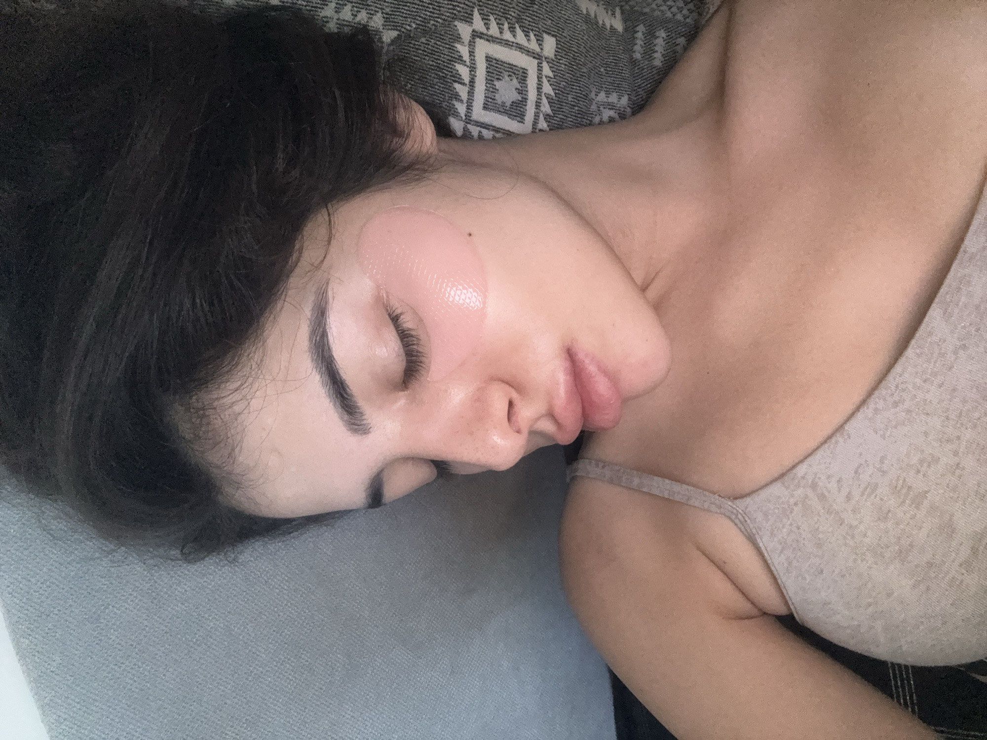 This is a close-up, top-down, eye-level shot of a young woman lying down, wearing under-eye patches and a beige tank top. She has dark hair and her eyes are closed. The background consists of a textured grey surface on the left and a patterned grey and white blanket on the right. There is no discernible location in the city of Plovdiv, Bulgaria.