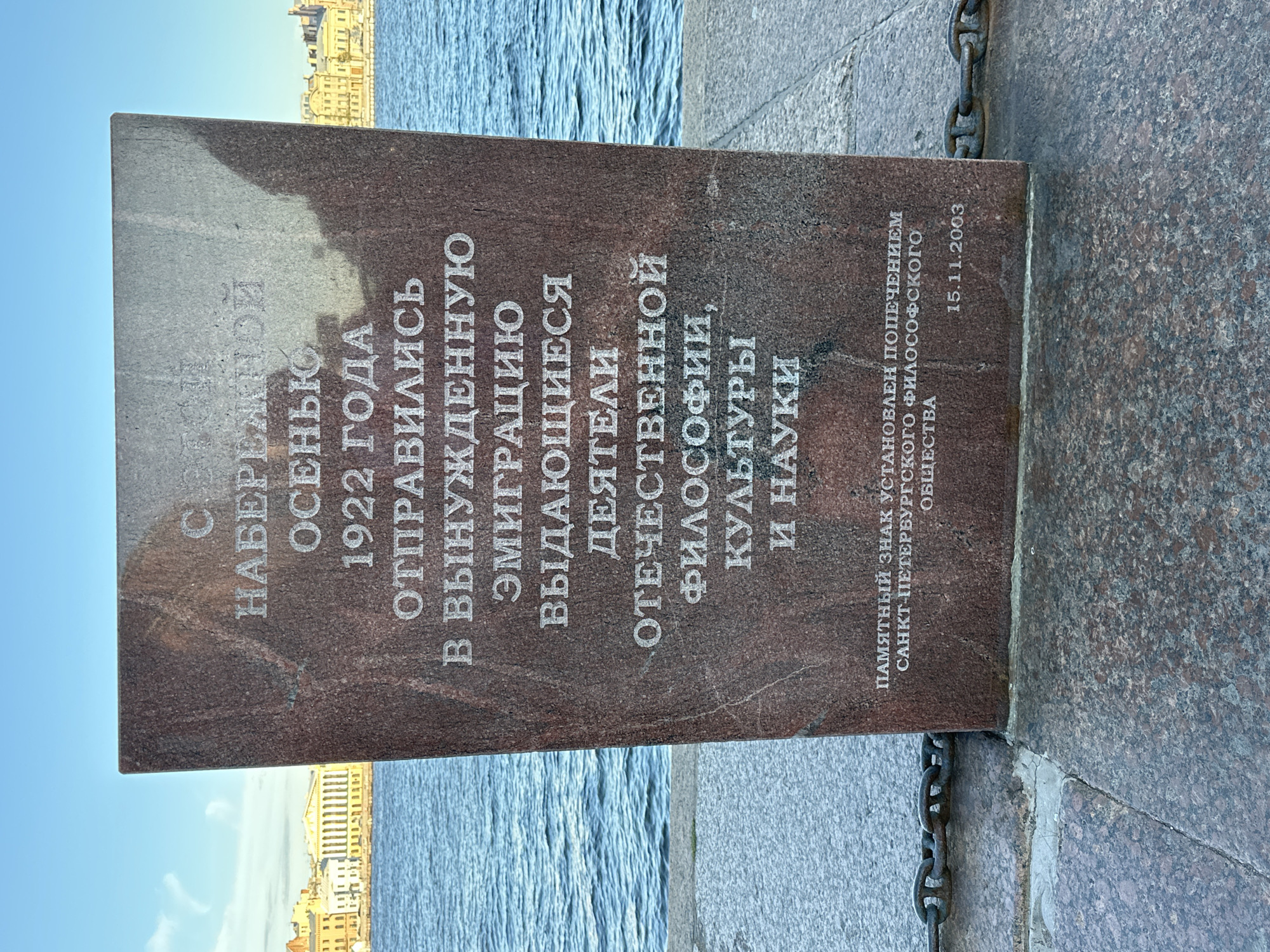 This image shows a vertical granite monument with inscriptions in Russian, set against a backdrop of a calm body of water and distant buildings with classical architecture. The monument is positioned on a paved walkway, with a heavy chain visible in the foreground. The sky is a clear blue, suggesting fair weather.
The inscription on the monument commemorates an event in the autumn of 1922, when distinguished figures from Russian philosophy, culture, and science embarked on forced emigration. It also states that the memorial sign was established by the patronage of the Saint Petersburg Philosophical Society on November 15, 2003. The text is carved into the reddish-brown granite, with the letters appearing lighter than the stone.
The scene likely takes place in Saint Petersburg, Russia, given the mention of the Saint Petersburg Philosophical Society and the architectural style of the distant buildings. The monument itself is the primary subject, serving as a historical marker for a significant period of emigration of Russian intellectuals. The overall mood conveyed is somber and reflective, acknowledging a loss of talent and intellectual heritage. The lighting suggests it is daytime.