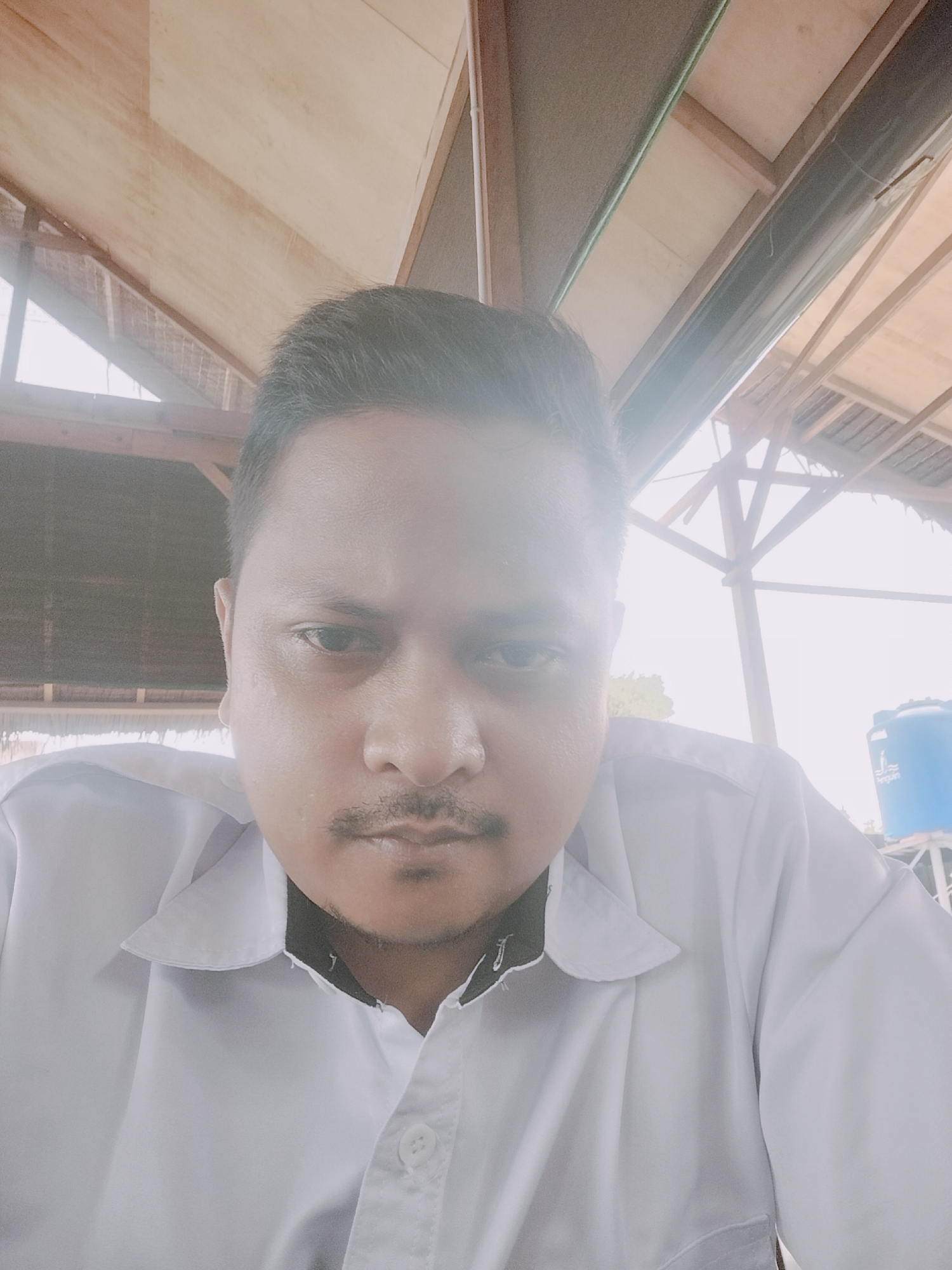 A medium shot, selfie-style photograph captures a man from the chest up. The man is wearing a white collared shirt with black trim around the collar. His hair is dark and neatly combed. He has a mustache and a serious expression. The background appears to be an outdoor setting with a wooden roof structure. To the right, a large blue water tank with the word "PENGUIN" is visible. The overall lighting is bright, suggesting it's daytime. The image is likely taken in Lhokseumawe, Indonesia, given the presence of the "PENGUIN" water tank, a common brand in that region.