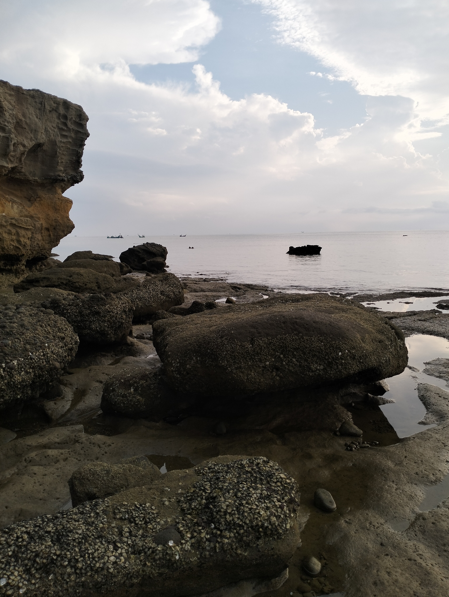 The image shows a rocky coastline in Lhokmanee, Indonesia. Large, dark, barnacle-covered rocks dominate the foreground, with pools of water reflecting the sky. A rock cliff is on the left. In the distance, the calm, grey ocean meets the overcast sky with boats visible. A rock formation is also visible in the ocean.