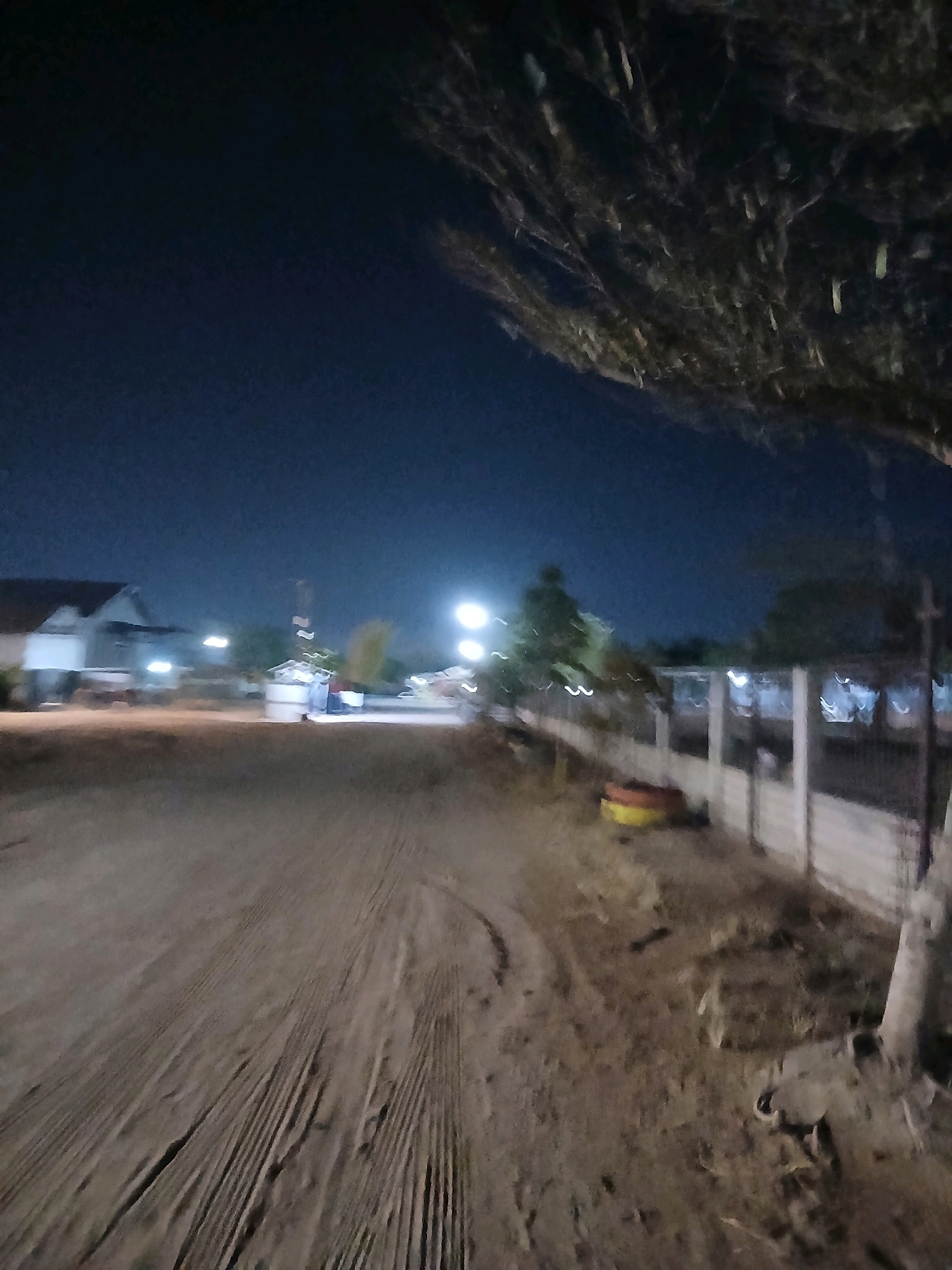 A dirt road stretches into the distance under a dark night sky, with branches of a tree visible in the upper right corner. The scene appears to be on the outskirts of Pangkalpinang, Indonesia, possibly in a rural or developing area. In the background, a building with a dark roof is illuminated by artificial lights, suggesting it might be a house or a small establishment. Further along the road, more lights are visible, possibly from streetlights or other buildings, creating a blurred effect due to motion or low light conditions. A fence runs along the right side of the road, with some objects of unclear nature situated near it. The ground is rough and uneven, with visible tire tracks, indicating vehicle activity. The overall impression is one of a quiet, dimly lit, and somewhat unkempt environment at night.