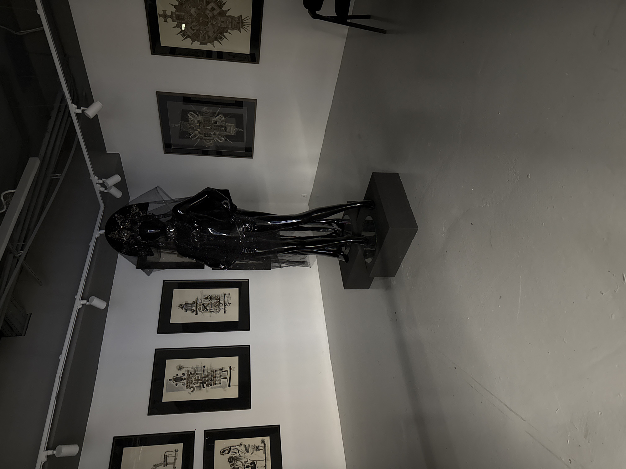 The image depicts an indoor space, possibly an art gallery, in Unknown, Unknown. On a white wall are hung several framed black-and-white drawings. To the right, a dark sculpture, potentially made of plastic or a similar material, stands on a dark base. Track lighting is visible near the ceiling. The floor and adjacent wall are gray, and the scene is illuminated by the gallery's lighting.
