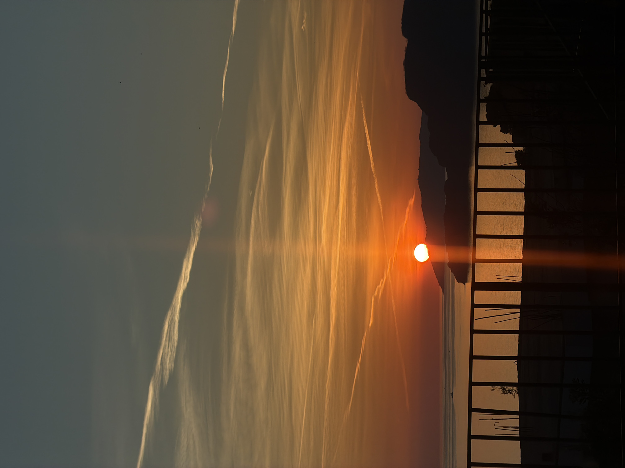 The image depicts a sunset over the Adriatic Sea, viewed from Sveti Stefan, Montenegro. The sun, a bright orange orb, is partially obscured by a mountain silhouette in the distance. Jet streams mark the sky. A dark, rectangular grid, possibly a balcony railing, frames the right side of the image. The sea reflects the sunset's orange light. The sky fades from a dark blue on the left to orange toward the horizon.