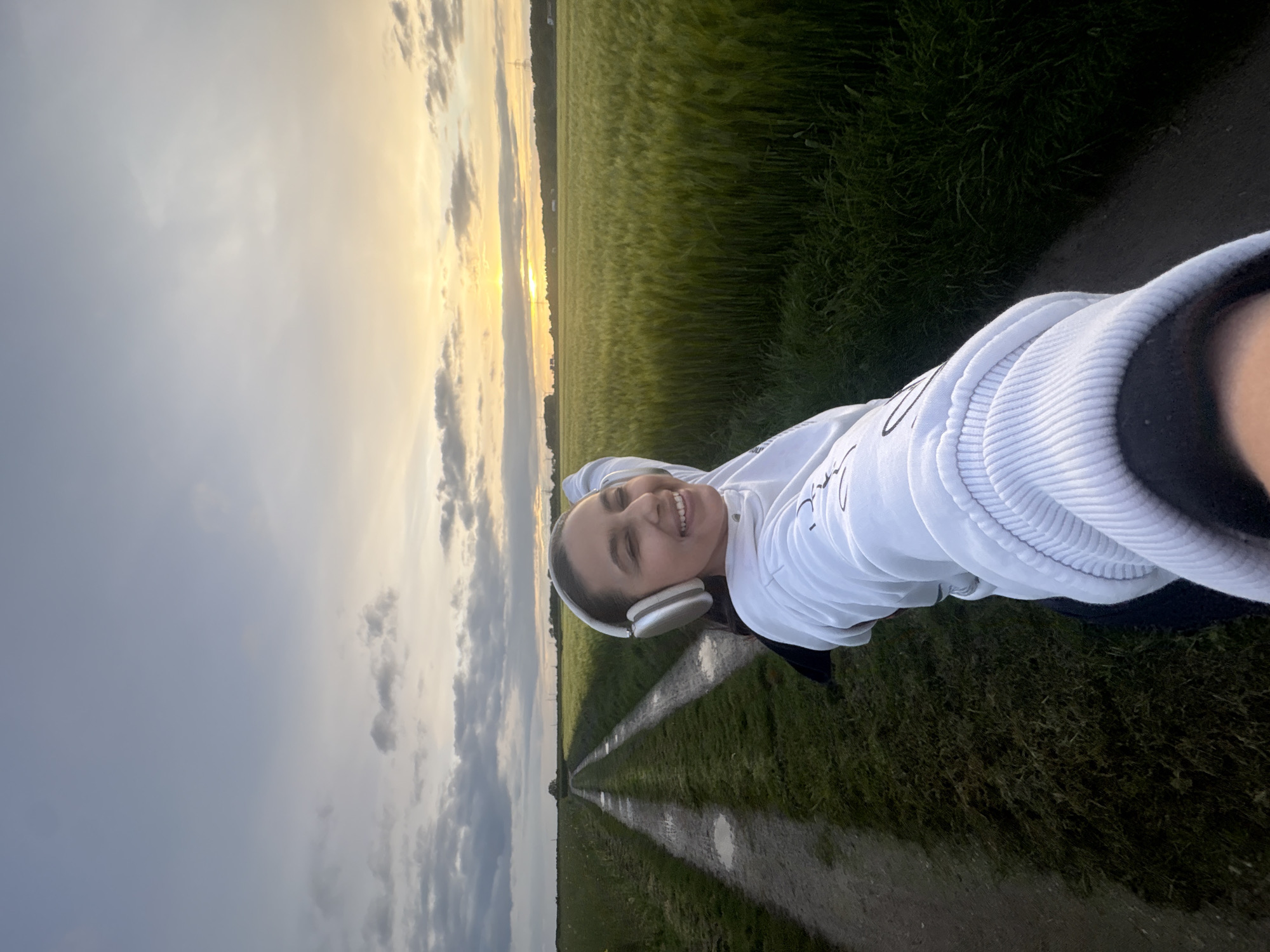 A woman with dark hair and fair skin is positioned in the foreground, wearing a white sweatshirt and white headphones, smiling, and looking at the camera. She appears to be taking a selfie. The setting appears to be a rural area in or near Mönchengladbach, Germany. She is standing on a dirt path running between fields of green grass. The sky overhead transitions from blue-gray with clouds to an area of golden light on the horizon. The dirt path has puddles of water.