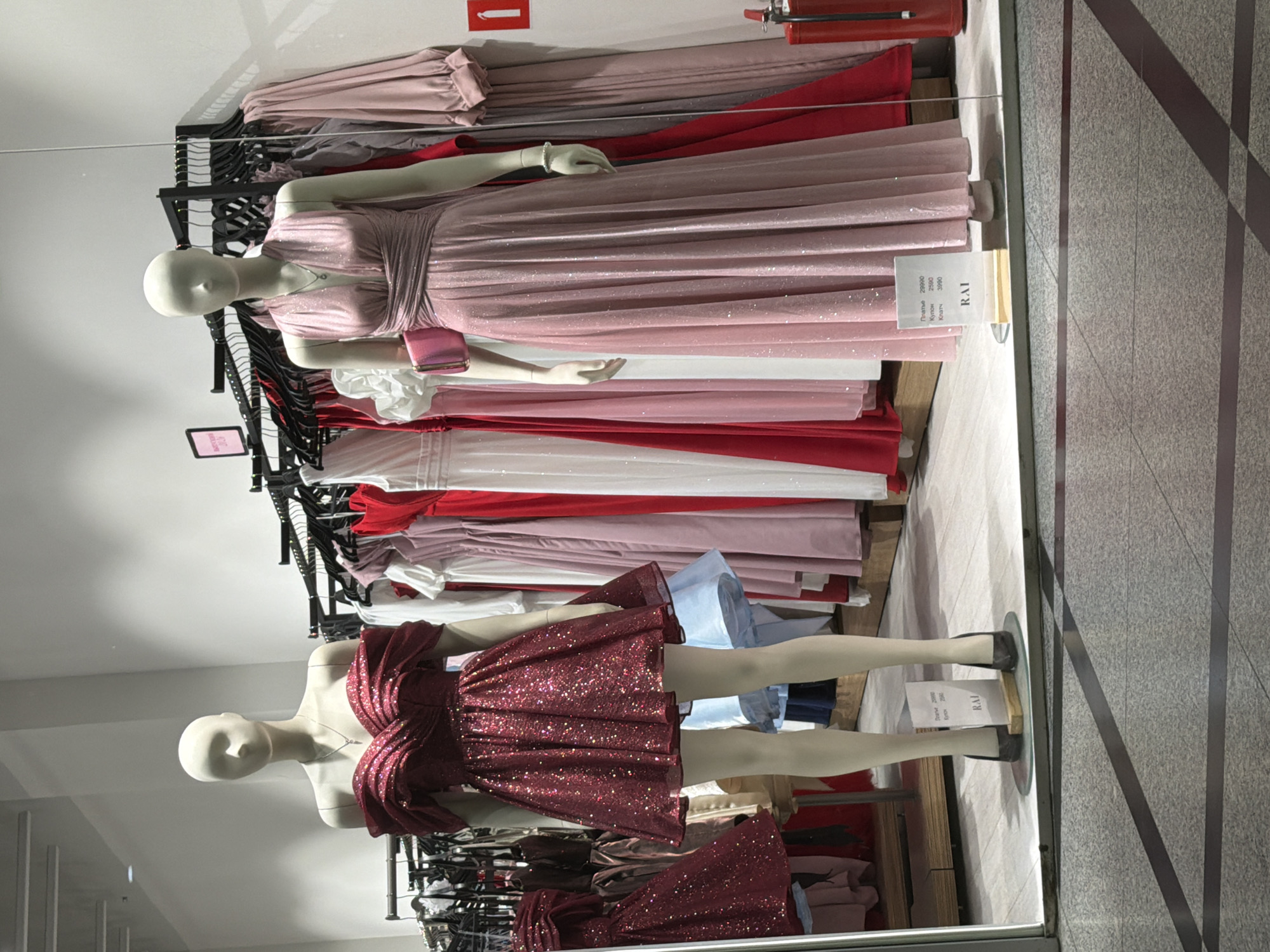 The image shows a shop window display featuring mannequins dressed in evening wear. The location appears to be a retail space, possibly in Yekaterinburg, Russia, based on the Cyrillic text on the price tags. The foreground features two headless mannequins, one wearing a short, red, sparkly dress and the other wearing a long, pink, sparkly dress with a matching handbag. Behind the mannequins are racks filled with dresses in various shades of pink, red, and white. A fire extinguisher is visible mounted on the wall near the top of the frame. The floor outside the window is a gray tile with dark stripes.