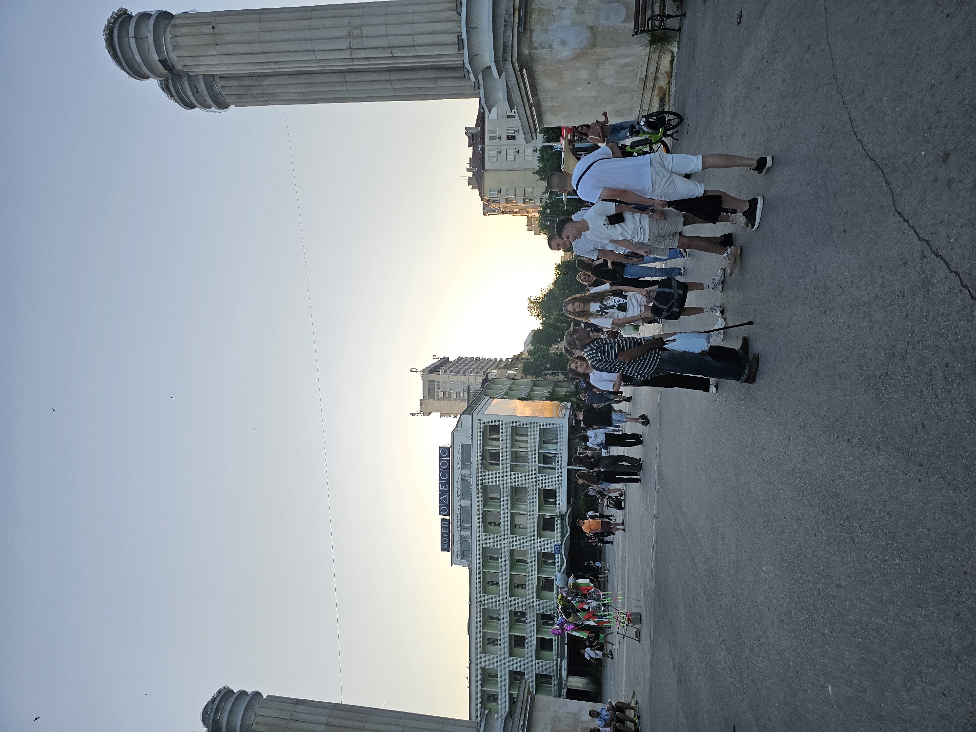 An outdoor scene captures a street area with a hotel in the background, two large columns on the right, and several people walking and standing. The sky is a pale blue with the sun setting, casting a warm glow on the buildings and the scene. A hotel sign with Cyrillic lettering, "ОДЕССОС", is visible above the hotel. People are dressed in casual summer clothing, and some are carrying bags or holding balloons. One person is using a walking stick. A green bicycle is parked near the columns. The ground is paved with asphalt.