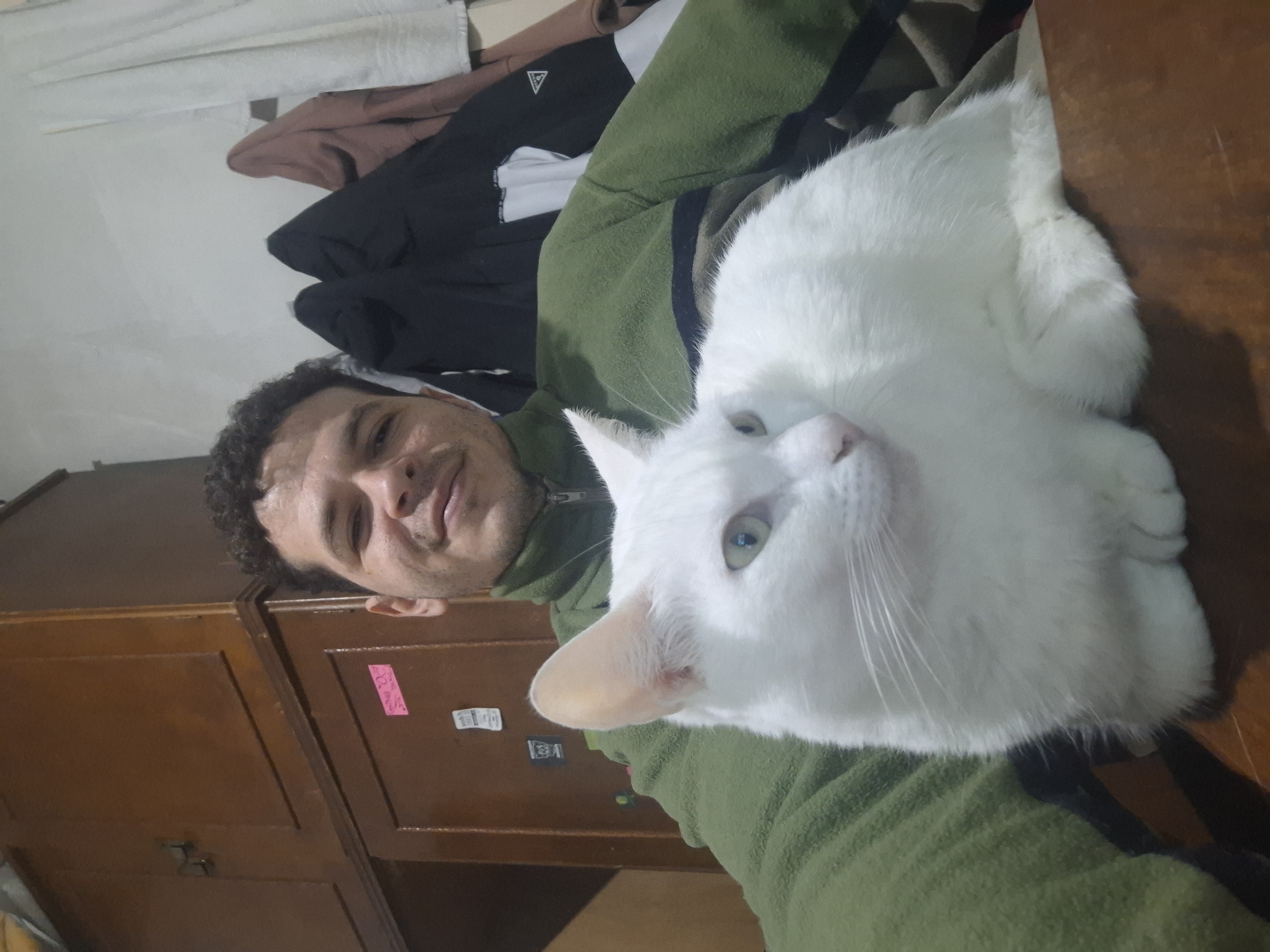 A man with curly dark hair and a light complexion is sitting, holding a white cat with green eyes. The man is wearing a green jacket. The cat is positioned on his lap, looking towards the left. In the background, there is a wooden cabinet with multiple doors and drawers. Various items, including jackets, are hanging on the wall behind the man and the cat. The scene appears to be indoors, possibly in a residential setting in Wilde, Argentina, given the context.