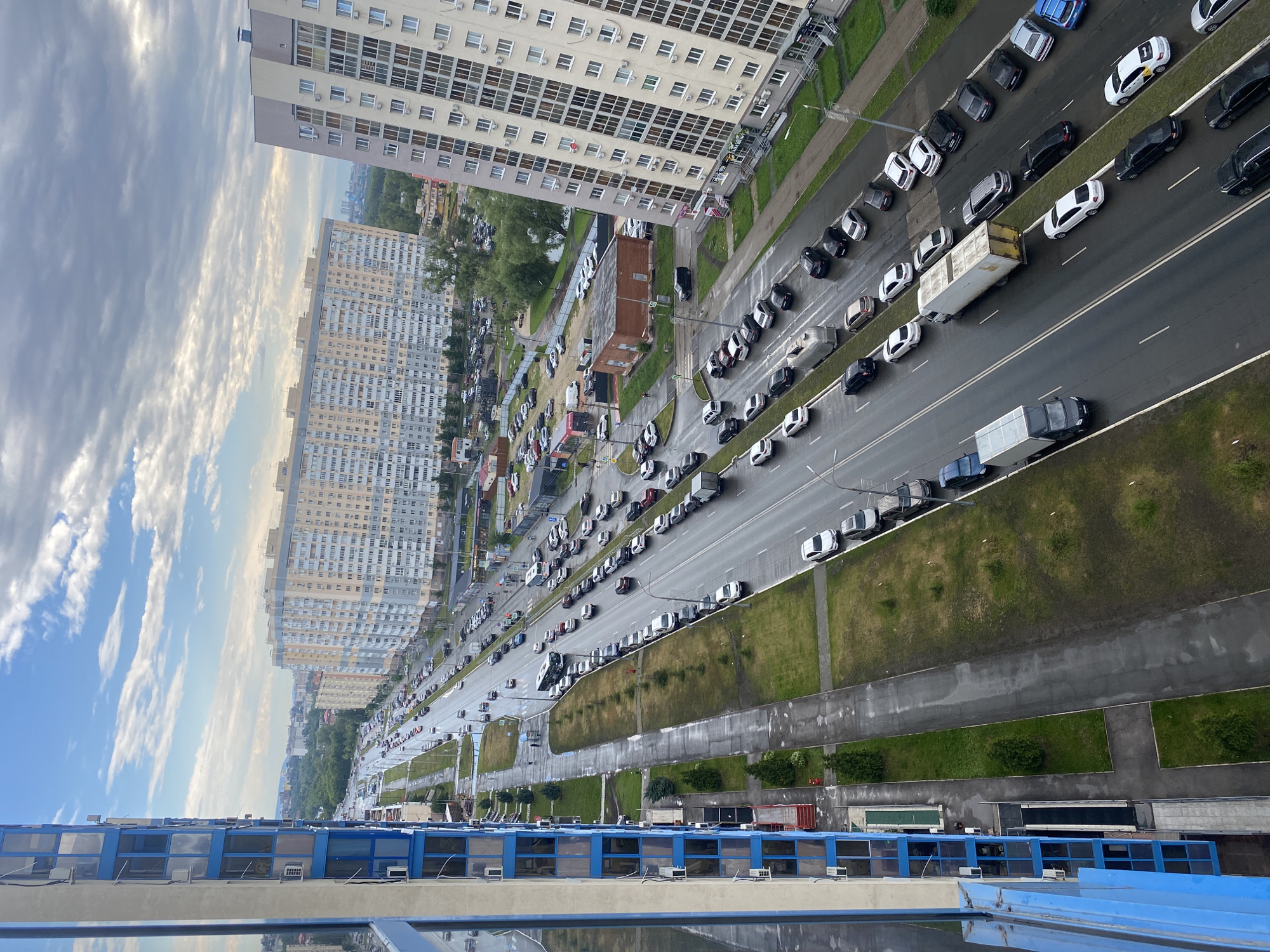 This is an aerial, high-angle, full shot of a multi-lane road in Novokuybyshevsk, Russia, with significant traffic. The road runs through a residential area characterized by several large apartment buildings. On the left side of the image, a modern, blue and grey apartment building with many windows is prominently featured. Across the street, on the right, are two other large, light-colored apartment buildings. The road itself is wet, suggesting recent rain, and has lane markings. Numerous cars, including sedans, SUVs, and at least two trucks (one a large white trailer truck), are present in both directions of traffic and parked along the roadside. Green grass and some trees line the road and sidewalks. In the background, more buildings and trees are visible, extending towards a hazy horizon. The sky is partly cloudy with a mix of blue patches and grey clouds. A few pedestrians can be seen on the sidewalks.