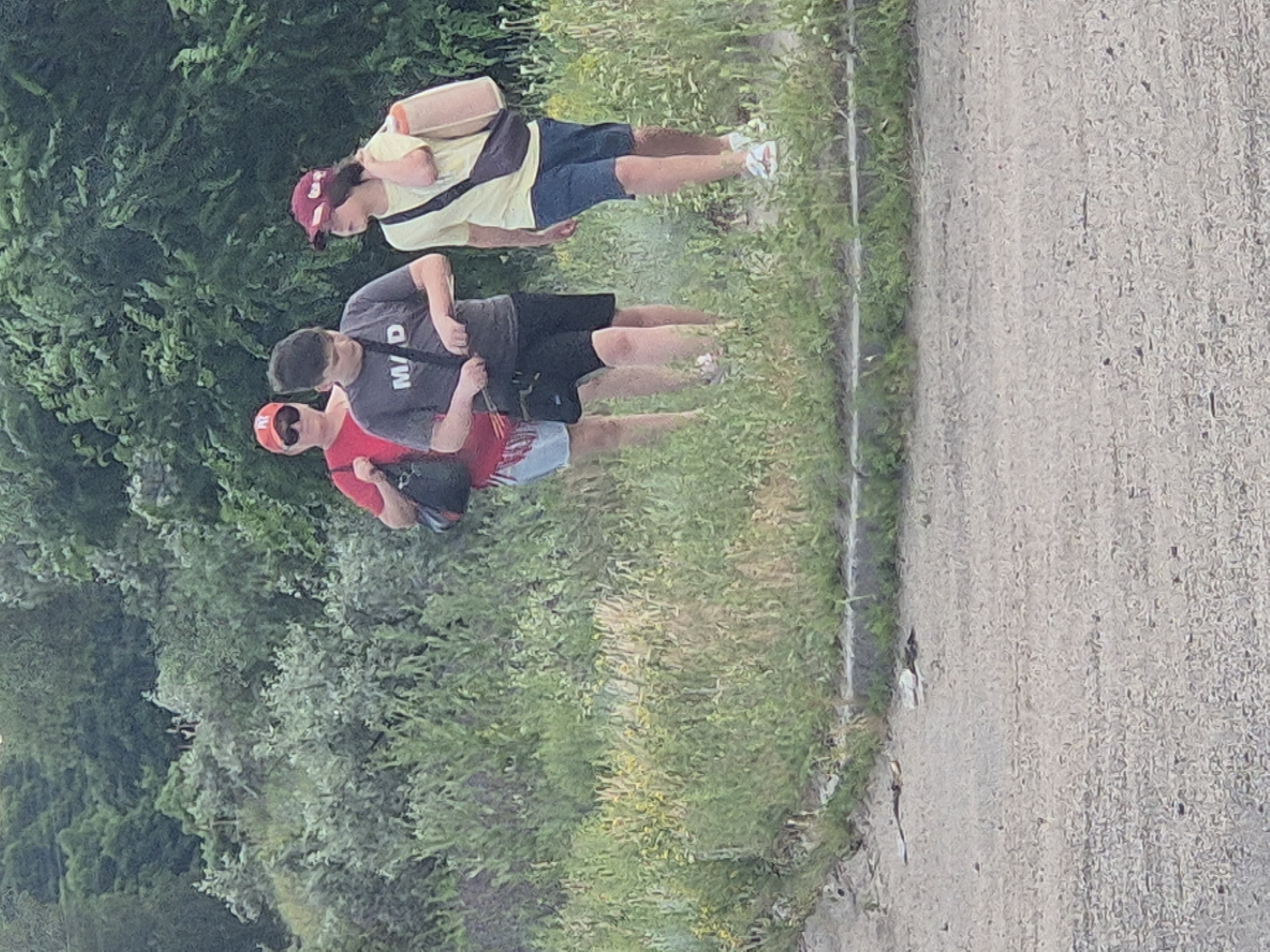 A medium shot captures three individuals walking on a dirt path alongside a grassy embankment. The person on the left, wearing a red t-shirt and a red cap, carries a black bag. In the center, a person in a gray t-shirt with the letters "M/D" printed on it, and black shorts, walks with a black strap across their body. To the right, a person in a yellow t-shirt and blue shorts, also wearing a red cap, carries a tan bag slung over their shoulder. The background is filled with lush green trees and vegetation. The scene appears to be outdoors, possibly in a park or natural area. The image does not provide specific details about the city of Pidhaitsi, Ukraine.