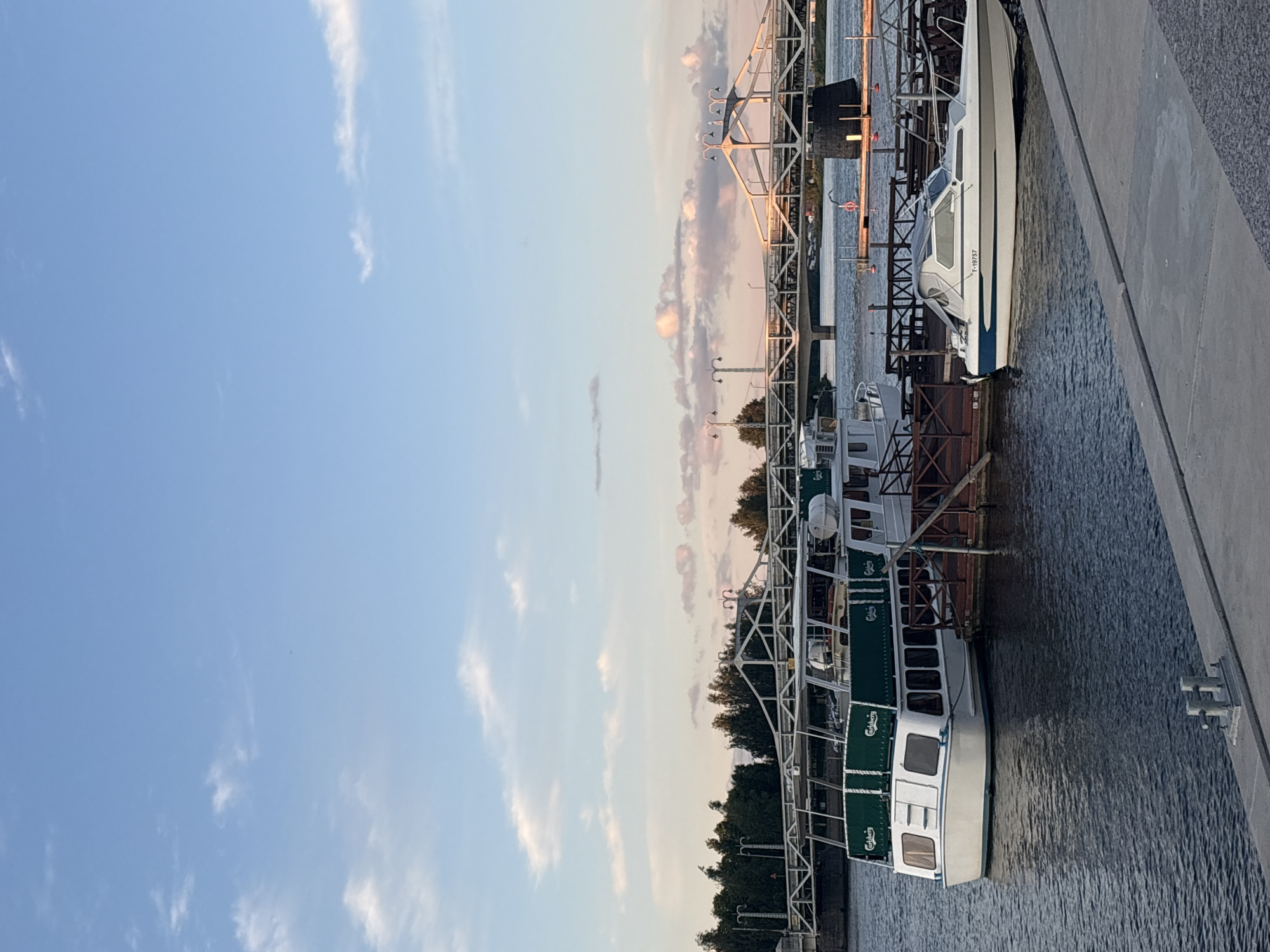 The image depicts a waterfront scene in Pori, Finland, likely during the late afternoon or early evening, with the sun casting a warm light. A large, white boat with green awnings displaying the "Carlsberg" logo is docked to the left. To its right, a smaller, sleek white motorboat with a blue stripe and registration number "T-19757" is also docked. Both boats are floating on dark blue water.
In the background, a white truss bridge spans across a body of water, with trees lining the far bank. The sky is a clear blue with scattered, soft pink and white clouds. The scene appears tranquil, suggesting a peaceful day at the harbor. The concrete embankment in the foreground provides a stable viewing platform.