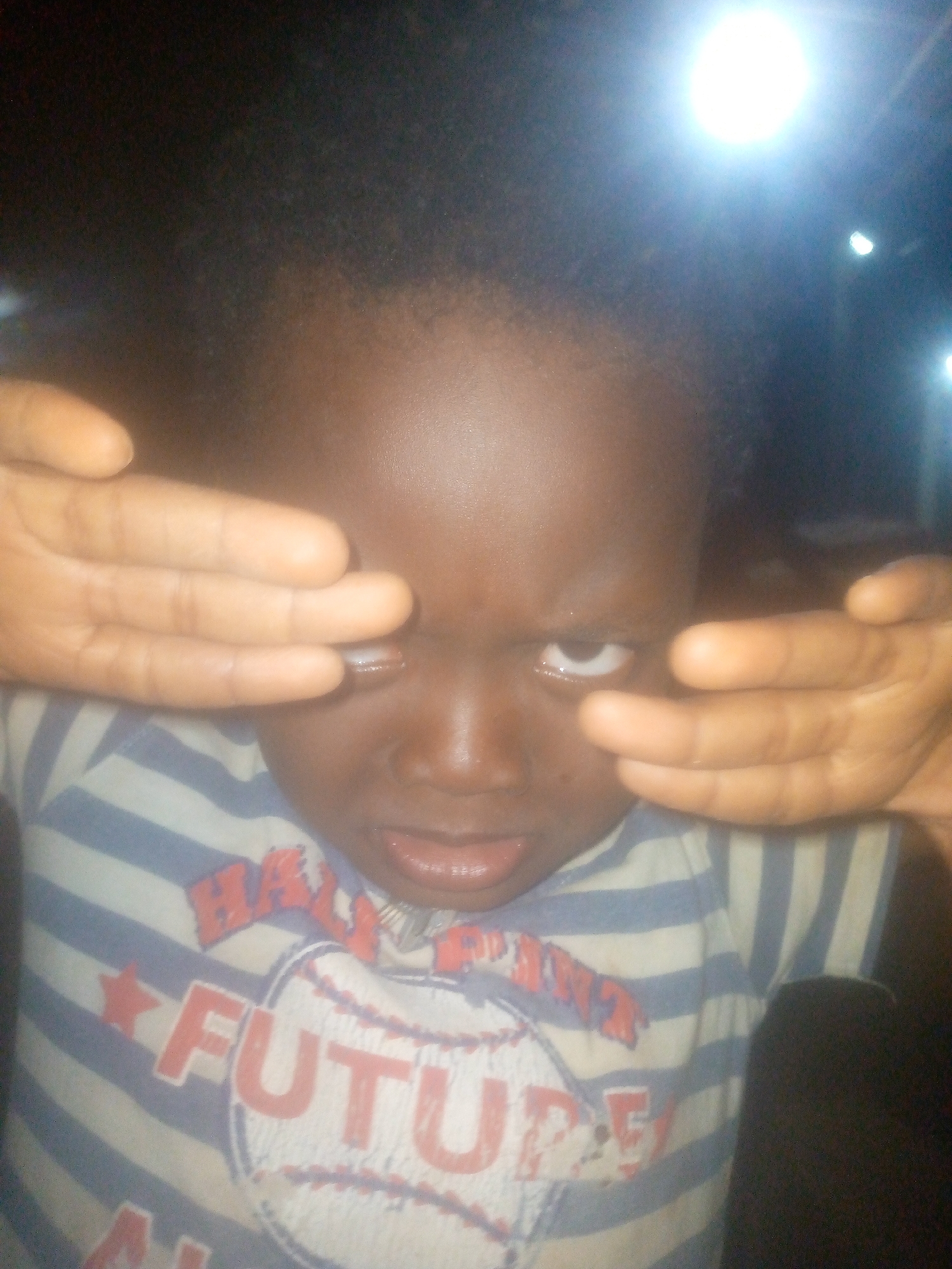 A close-up portrait features a young child, likely a toddler, with dark skin and dark, curly hair. The child is positioned centrally in the frame, from the chest up. Both of their hands are raised, palms facing outwards, covering their eyes as if shielding them from a bright light or engaging in a game of peek-a-boo. Through the gaps between their fingers, the child's eyes are visible, looking slightly upwards with an intense or curious gaze. Their mouth is slightly open, revealing a portion of the tongue.
The child is wearing a horizontally striped t-shirt with alternating light blue and white stripes. On the front of the shirt, a prominent graphic depicts a white baseball with red stitching. Overlaid on the baseball are bold red capital letters spelling out "FUTURE." Above "FUTURE," to the left, a red star is visible, and additional red text, partially obscured, appears to spell "BASEBALL."
The background is dark and out of focus, indicating a low-light environment, likely indoors. Several bright artificial light sources are visible in the upper half of the frame, with a particularly intense, circular white light dominating the top center-right, casting a significant glow and some lens flare. Two smaller, less intense lights are also present in the upper right. The strong backlighting and prominent artificial light sources suggest the image was captured at night or in a dimly lit room. The child's expression is ambiguous, hinting at playfulness, curiosity, or being momentarily overwhelmed by the bright lights.
