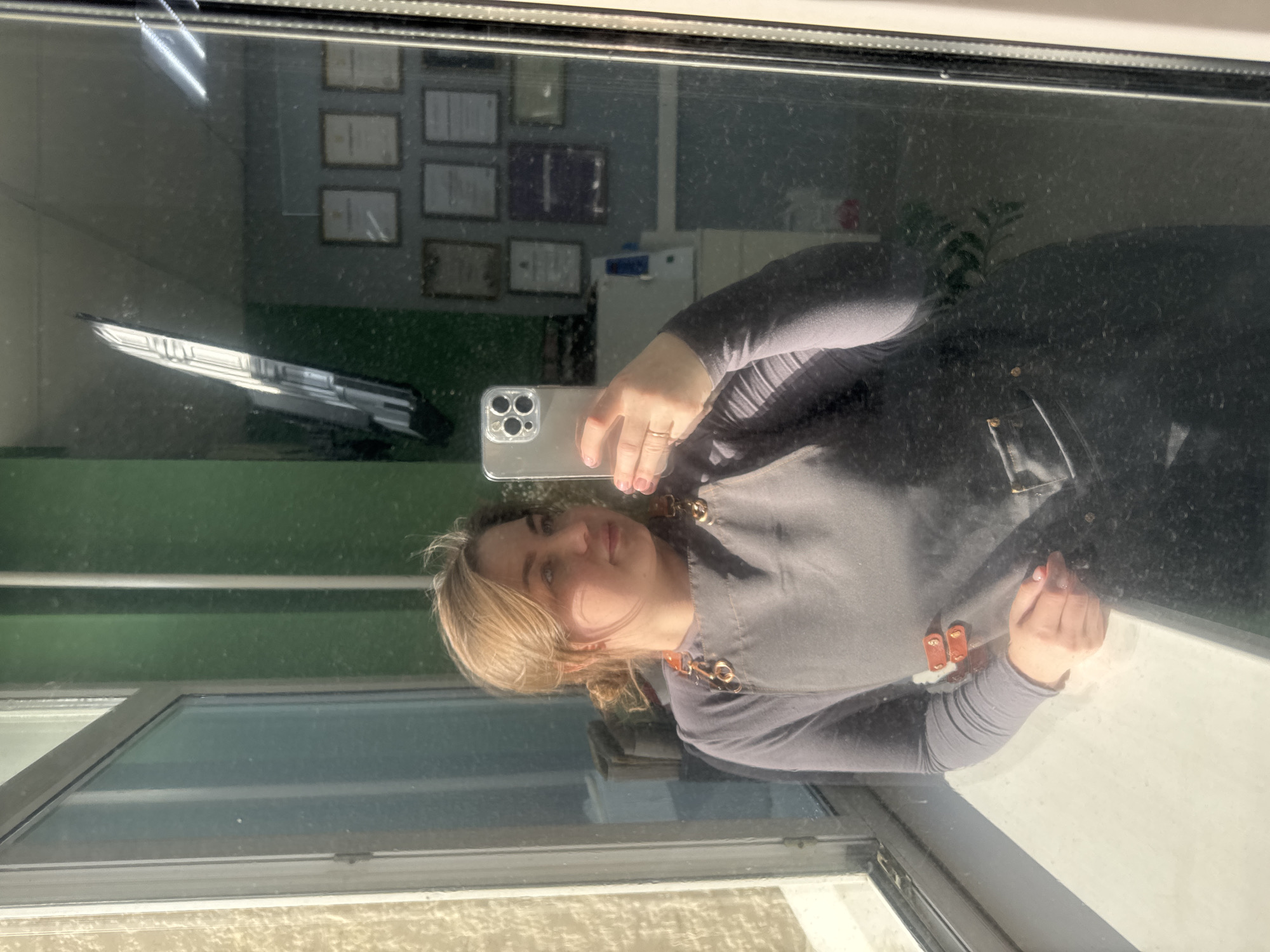 A woman with blonde hair is taking a selfie in a window, holding a silver iPhone 13. Her reflection is visible in the glass, showing her wearing a grey top and a black apron with brown leather straps. Reflected in the window, there are several framed certificates or diplomas hanging on a green wall. Also reflected are fluorescent lights and a white cabinet. The location and city are not possible to determine from the image, but it is stated in the prompt to be the city of Chaikovsky, Russia. The exterior of the window reveals a cream-colored wall and a window frame.