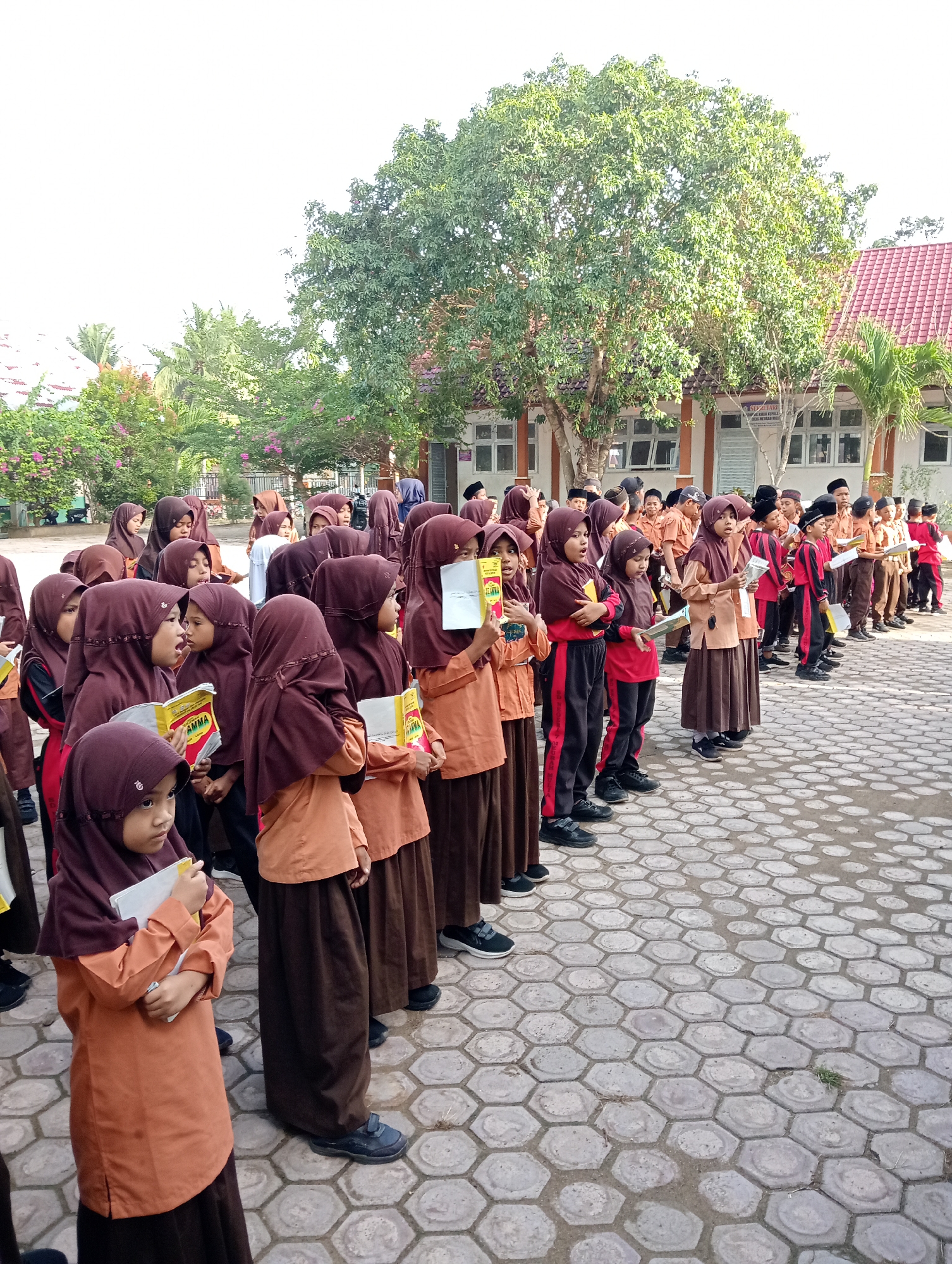 In Baratpayaitek, Indonesia, a group of school children, primarily girls wearing brown hijabs and orange tops with brown skirts, stand in formation on a paved, hexagonal-patterned courtyard. Some of the girls are holding books and appear to be listening or reciting. Behind them, more students and a building with red-tiled roofs are visible. The scene is outdoors, with trees providing shade and a bright sky overhead.