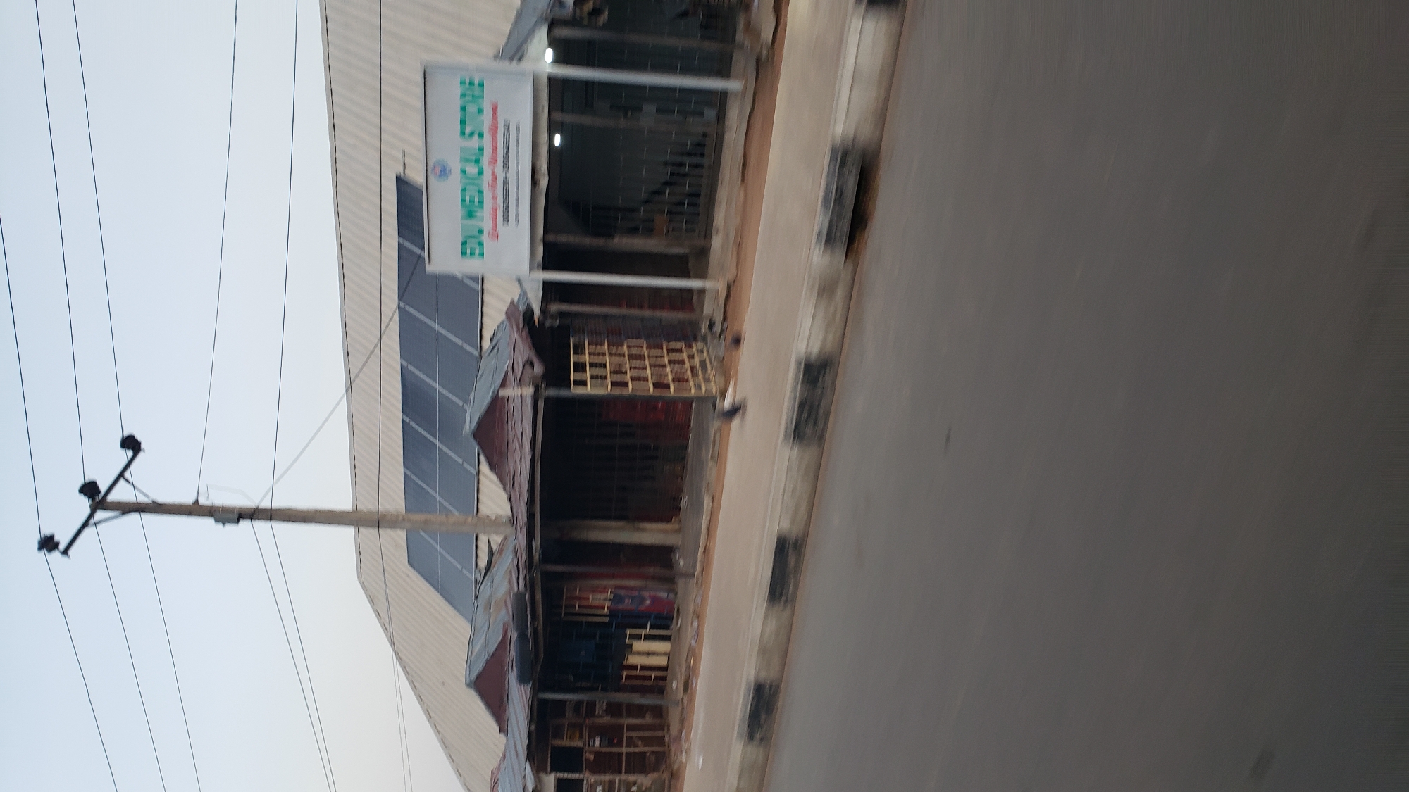 The image shows a street scene in Jalingo, Nigeria. A row of small shops with corrugated iron roofs line the sidewalk. One shop, labeled "EDU MEDICAL STORE," is visible with a white sign above its entrance. The buildings are simple, likely constructed with local materials. In the background, a large metal roof structure is partially visible, fitted with solar panels. A utility pole with electrical wires stands prominently in the foreground. The street in front of the shops appears to be paved. There are no people clearly visible in the frame, though some birds are on the sidewalk. The overall impression is of a typical urban commercial area in Nigeria.