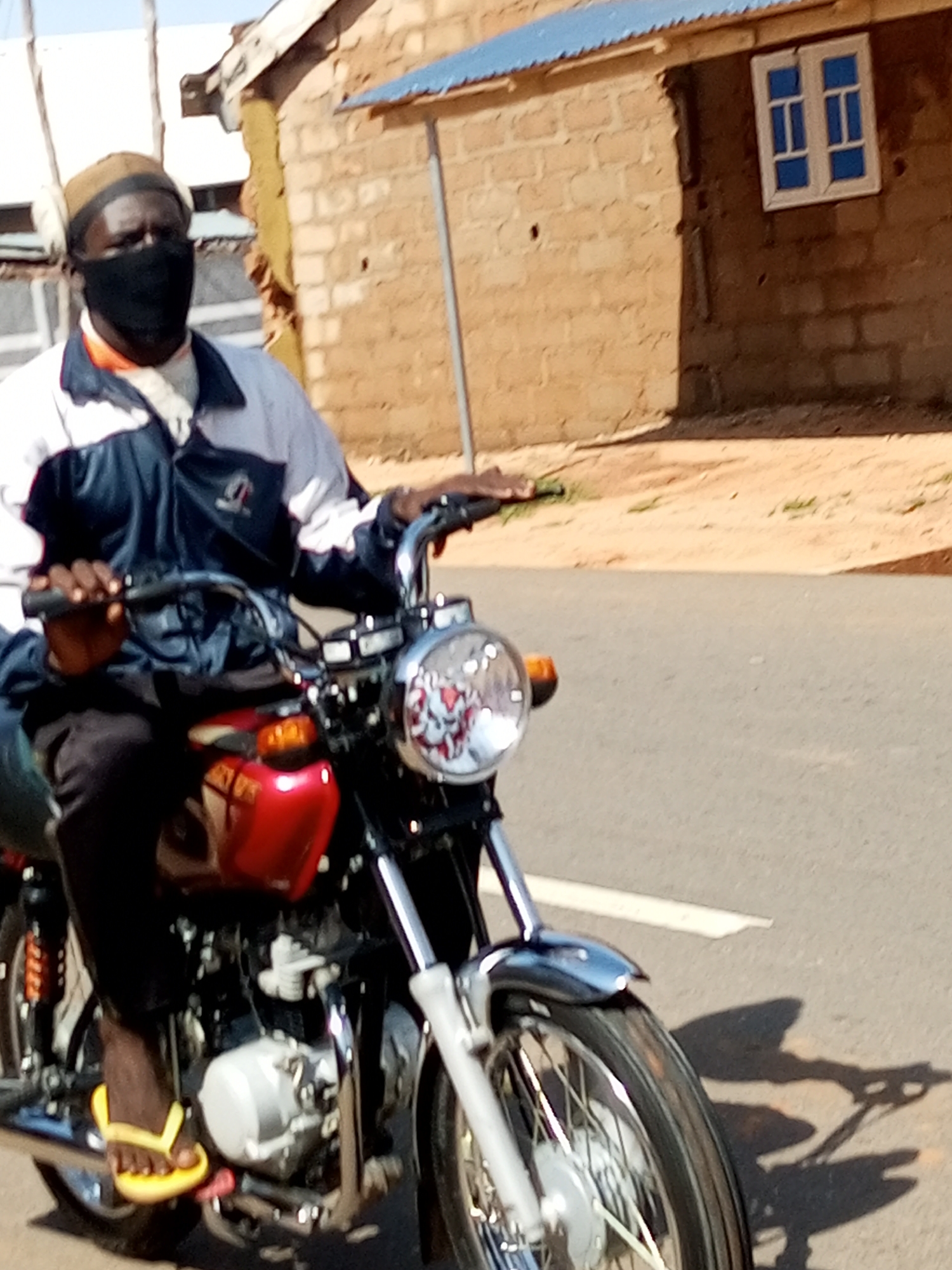 In Arawa, Nigeria, a man wearing a dark face mask and a light brown cap rides a red motorcycle on a paved road. He is dressed in a blue, white, and black jacket over an orange shirt. His left foot, wearing a yellow flip-flop, rests on the motorcycle's footrest. The motorcycle is a red and silver model with a prominent headlight. In the background, a beige brick building with a blue-roofed overhang and a small blue-paned window is visible, with dry, sandy ground in front of it. The lighting suggests it is daytime.