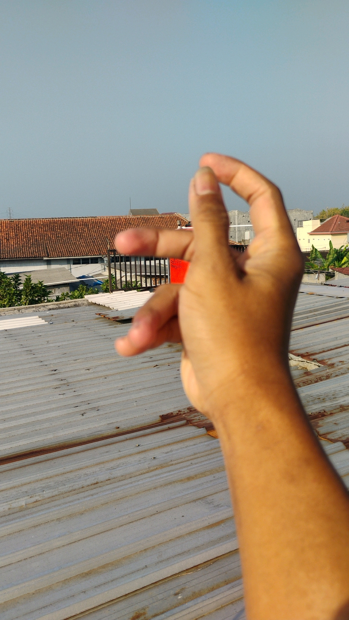 An outdoor, eye-level, close-up shot shows a right hand with tanned skin held up in the foreground. The fingers are partially closed, with the thumb and index finger forming a pinch gesture. The hand is positioned at an angle that suggests it is either reaching out or being photographed.
In the mid-ground, a metal roof with visible corrugated texture and some rust marks stretches across the frame. Behind the roof, there are buildings with terracotta-tiled roofs and white walls, characteristic of residential or commercial structures in Indonesia. A red rectangular object with some white text is visible behind the hand. Trees with green foliage are also present in the background.
The sky above is clear and a pale blue, indicating a daytime scene with bright sunlight. The overall impression is a casual snapshot taken from a rooftop in an urban or suburban environment, possibly in Gumpang, Indonesia, given the architectural style.