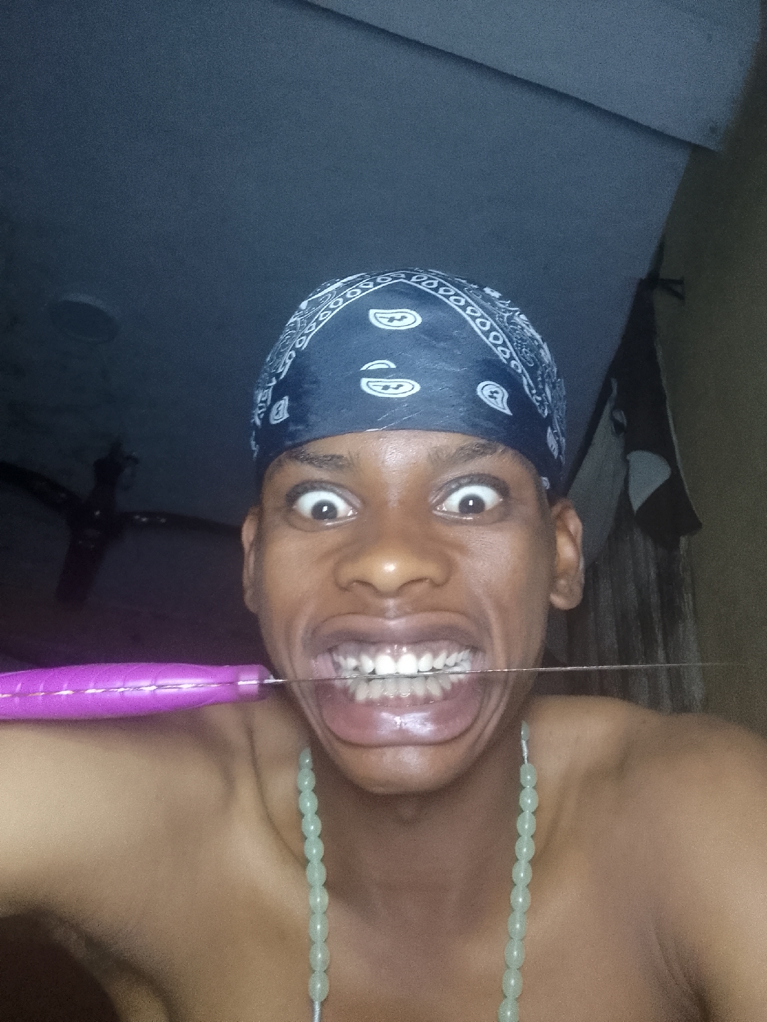A close-up indoor shot features a shirtless individual with a dark blue bandana on their head. The person's eyes are wide open, mouth open, and teeth bared. A thin metal object is held in the mouth, extending outwards. A green beaded necklace hangs around the person's neck. The location is Nkpor, Nigeria. There is a ceiling fan partially visible in the background.