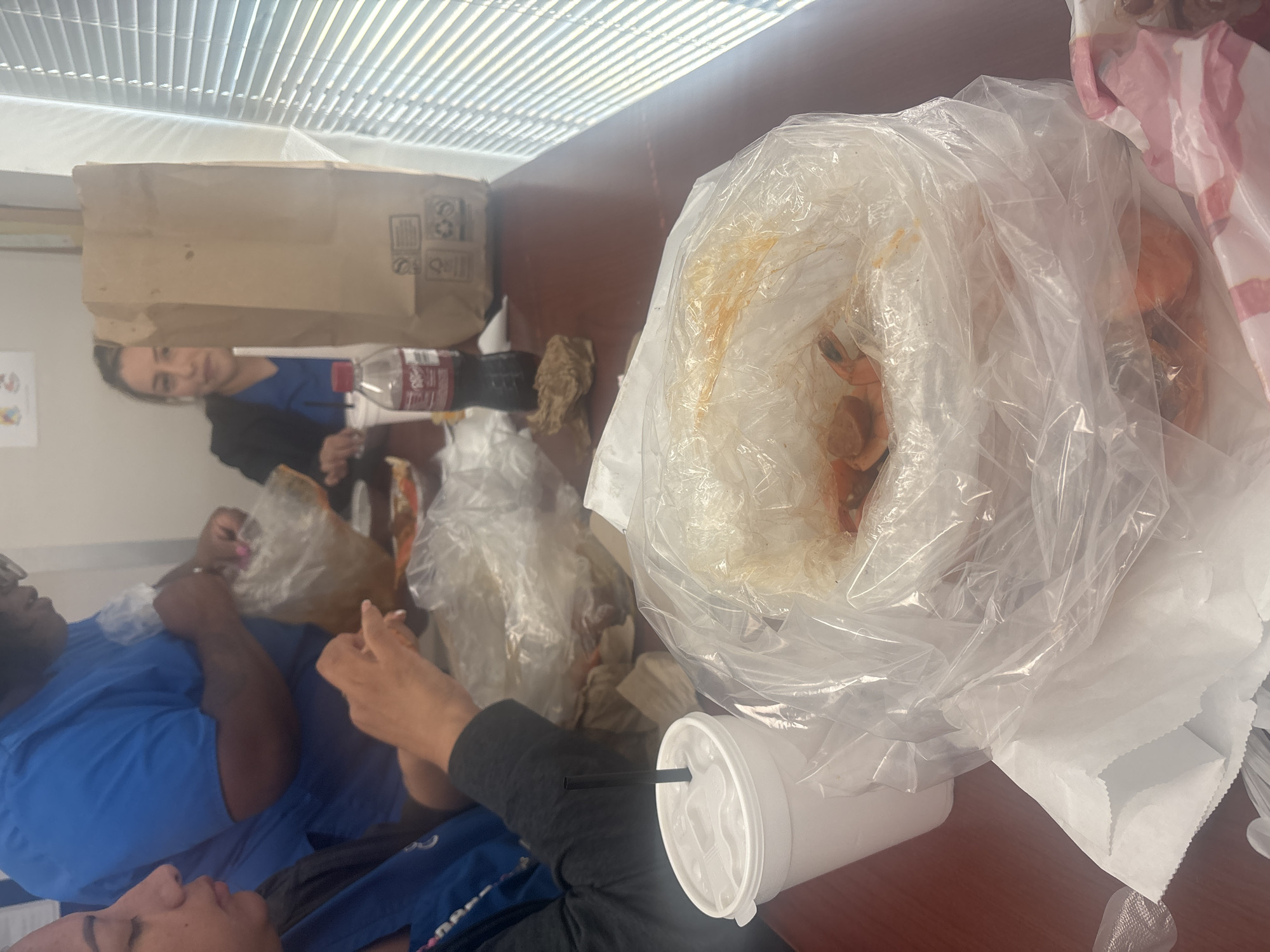 The image captures an indoor scene in Grand Prairie, Texas. A group of people are gathered around a table. One person wearing a blue shirt can be seen on the left side, holding a bag of food, another person wearing a black shirt with blue sleeves is seen further down. A woman with dark hair is sitting to the upper right of this person wearing a blue shirt, she is looking at the camera.
On the table are numerous clear plastic bags filled with food, likely seafood. There is a large white paper cup with a straw, a brown paper bag, and a bottle of Coca-Cola. The environment appears to be a casual dining or break room setting, with the blinds in the background indicating the presence of a window.