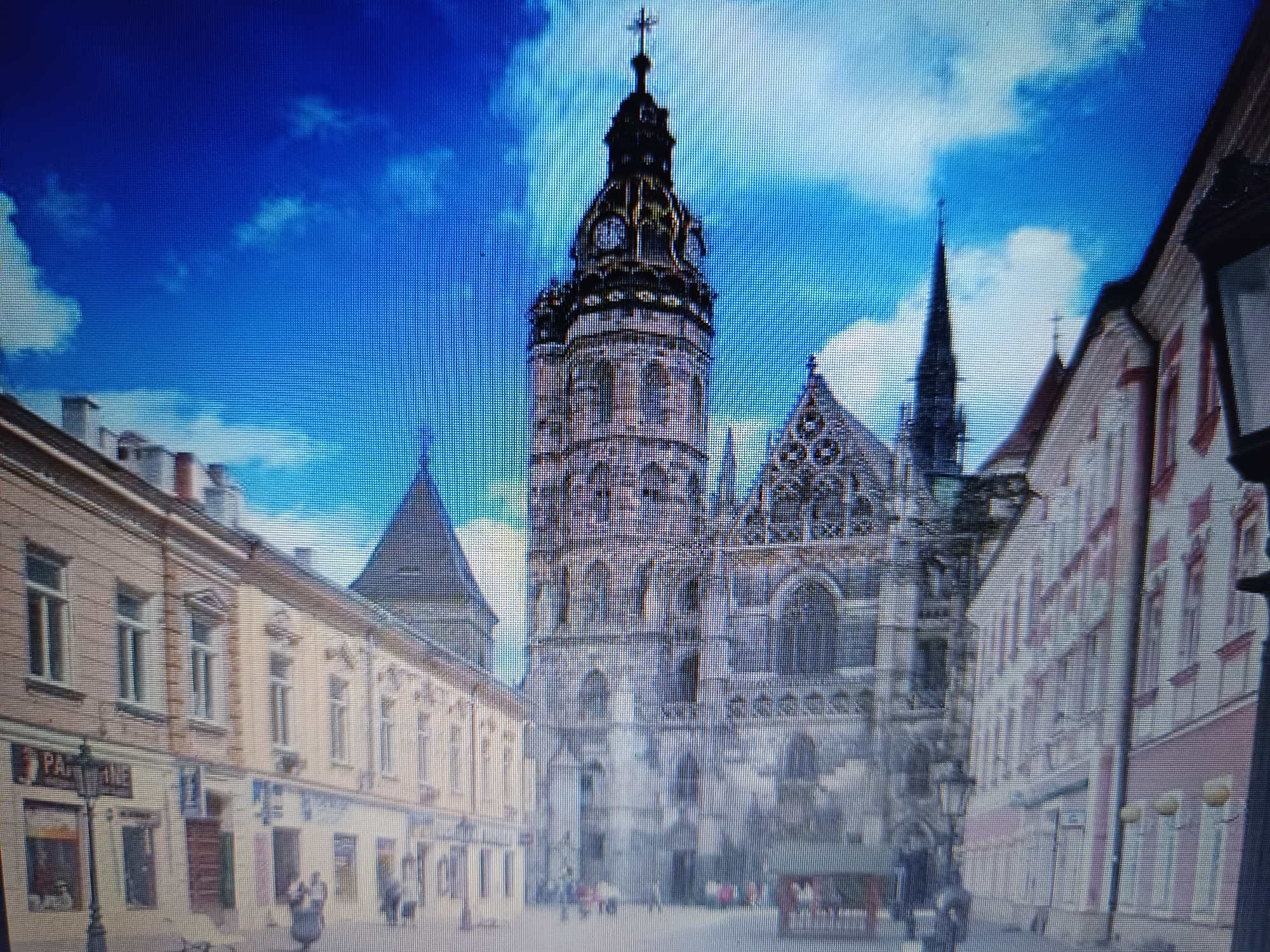 The image captures a street view in the city of Košice, Slovakia. In the center, St. Elizabeth's Cathedral, a prominent Gothic church, dominates the scene with its intricate architecture and tall spire topped with a cross. To the left of the cathedral, a series of light-colored buildings with traditional architectural features line the street, featuring multiple windows and balconies. A street lamp with a sign that reads "PAR" is visible in front of one of these buildings. On the right side of the image, another building with a pink facade and white trim stands. The street is paved, and in the foreground, a few people are present, some walking and others appearing to be interacting. The sky is a vibrant blue with white clouds, suggesting fair weather. The overall scene depicts a historic urban setting with significant architectural landmarks.