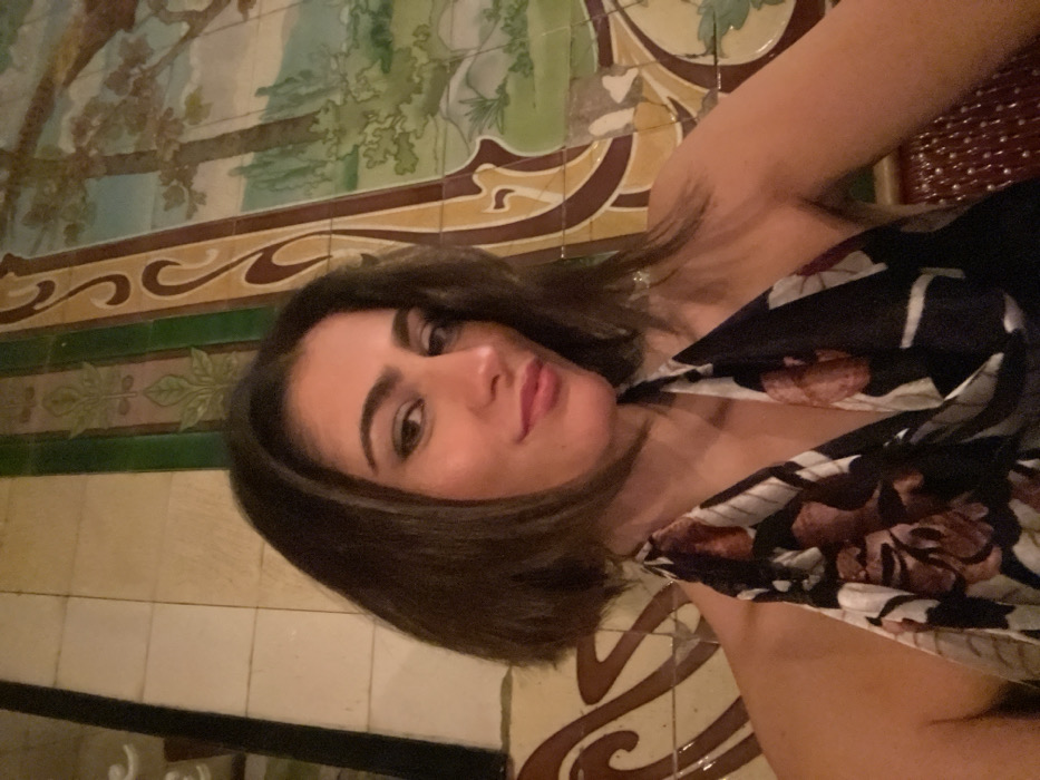 A brunette woman poses close to the camera. She has fair skin, dark eyebrows, and a pink lip. Her hair is short and dark. She wears a black dress with a floral pattern. The background consists of tiled walls in greens, browns, and whites with various patterns. The tiles appear vintage and decorative.
