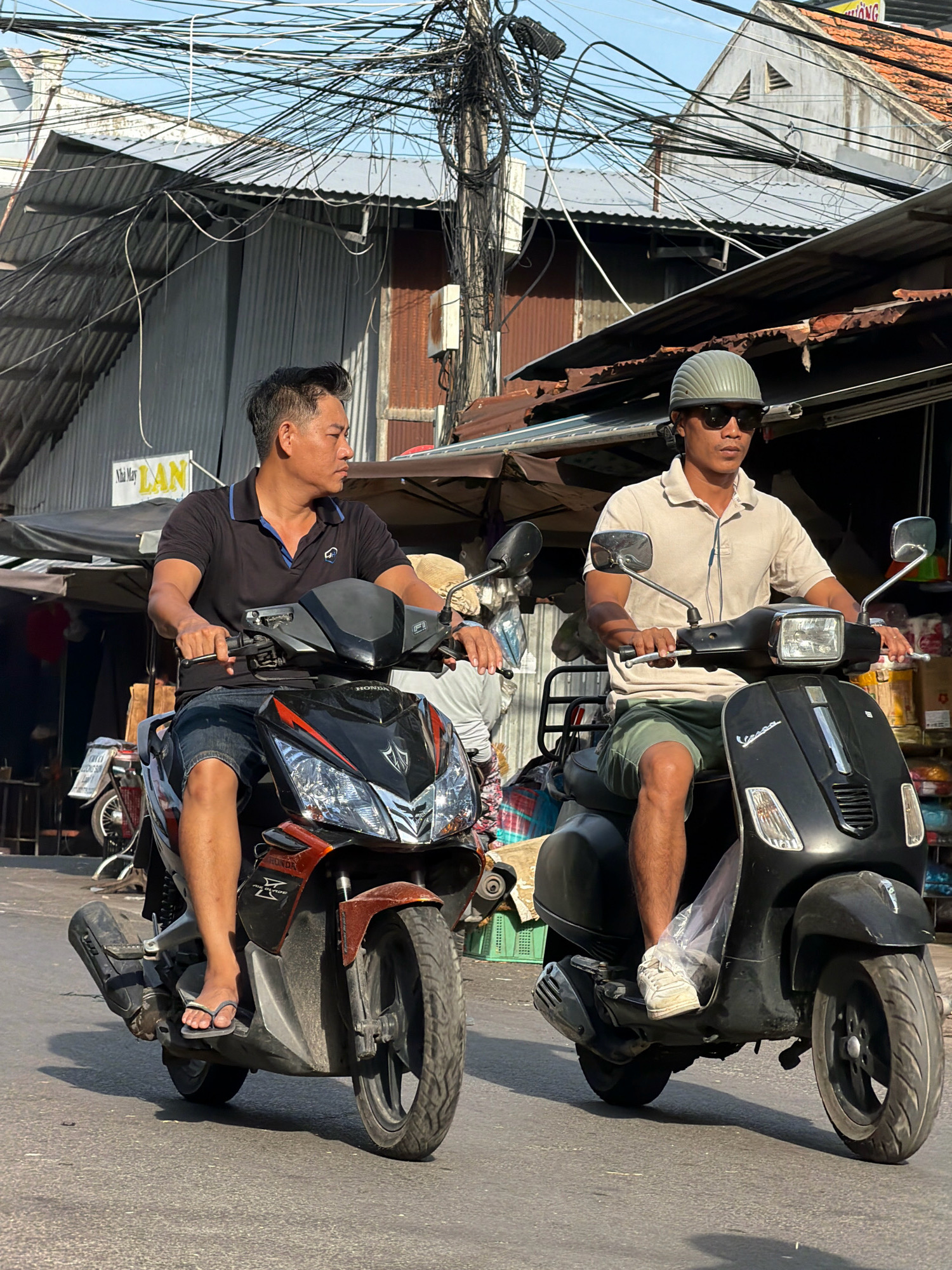 Two men ride motorcycles down a street in Nha Trang, Vietnam. The man on the left, wearing a black polo shirt and denim shorts, rides a black and orange Honda motorcycle. He is looking to the right. The man on the right, wearing a light-colored polo shirt and green shorts, rides a black Vespa scooter. He is wearing sunglasses and a light green helmet, and is looking forward. The street is lined with buildings that appear to be shops, with corrugated metal roofs and awnings. Overhead, a dense network of electrical wires crisscrosses the sky. The lighting suggests it is either early morning or late afternoon, with long shadows cast on the road.