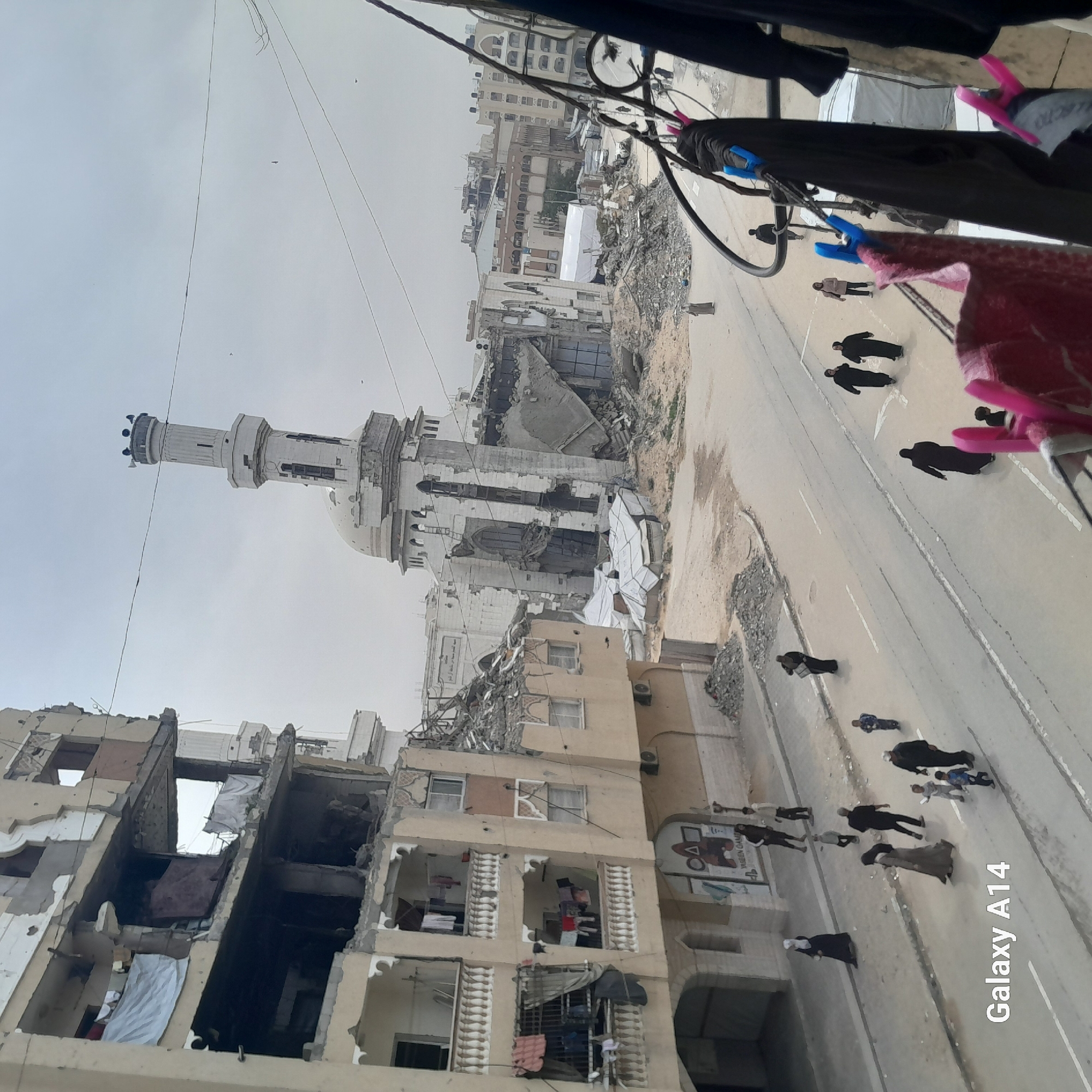 This image shows significant destruction to buildings, including a mosque with a damaged dome and minaret. The scene is likely in an urban environment, given the multiple buildings and road. A group of people are walking on the road, many of whom are wearing dark clothing. Debris is visible around the damaged buildings. Clothes are hanging out to dry in the upper right of the image. A phone imprint is in the corner of the image.