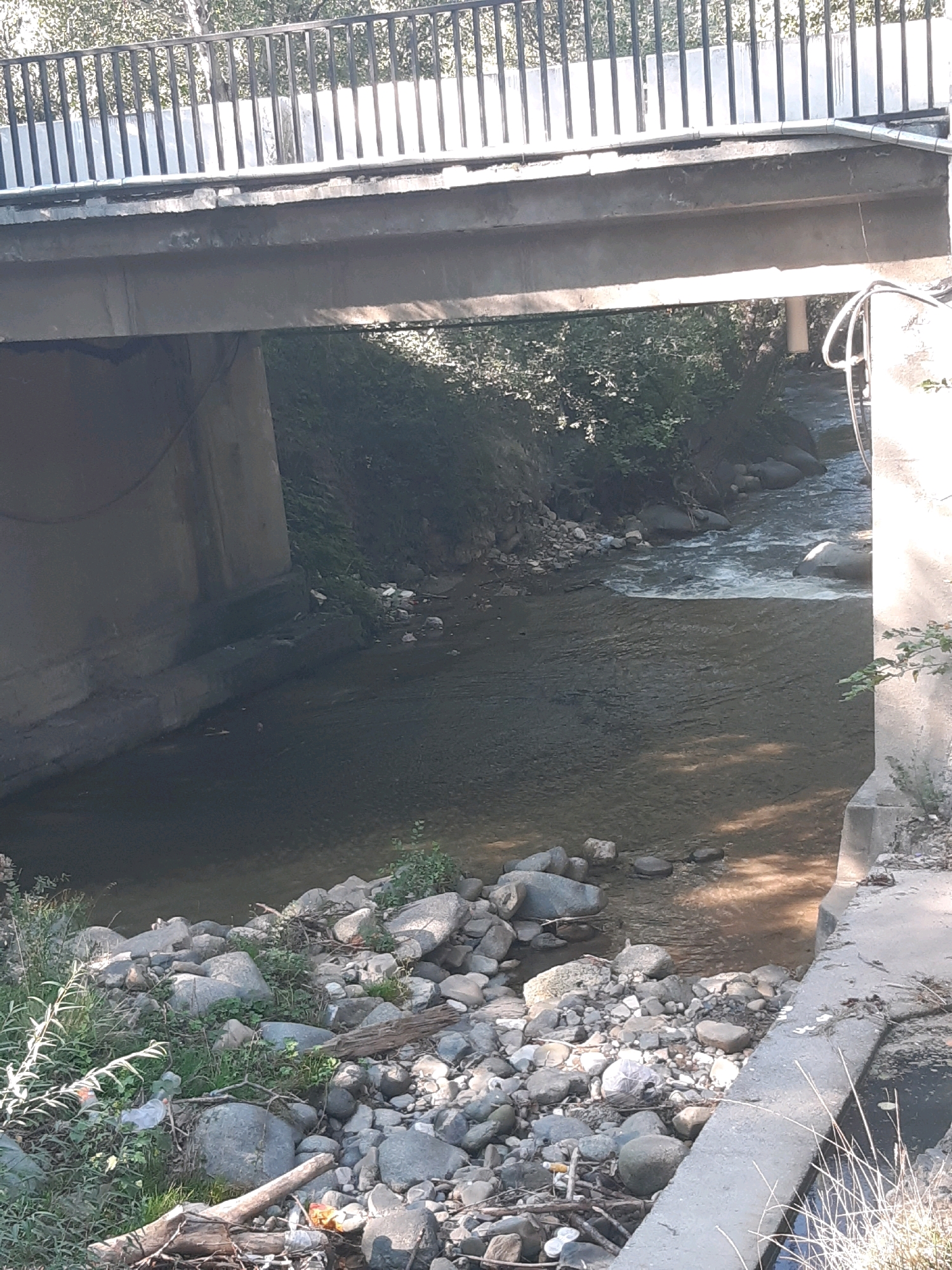 The image shows a concrete bridge spanning a shallow, rocky stream in Kavtiskhevi, Georgia. The bridge has a metal railing on top and concrete supports. The stream bed is filled with various sized rocks and pebbles, and some vegetation grows along its banks. There is evidence of some litter on the banks and in the water, including plastic bottles and wrappers. The water appears murky and reflects the sunlight in places. The surrounding environment is natural, with trees and foliage visible on the banks of the stream. The scene suggests a rural or semi-rural setting.