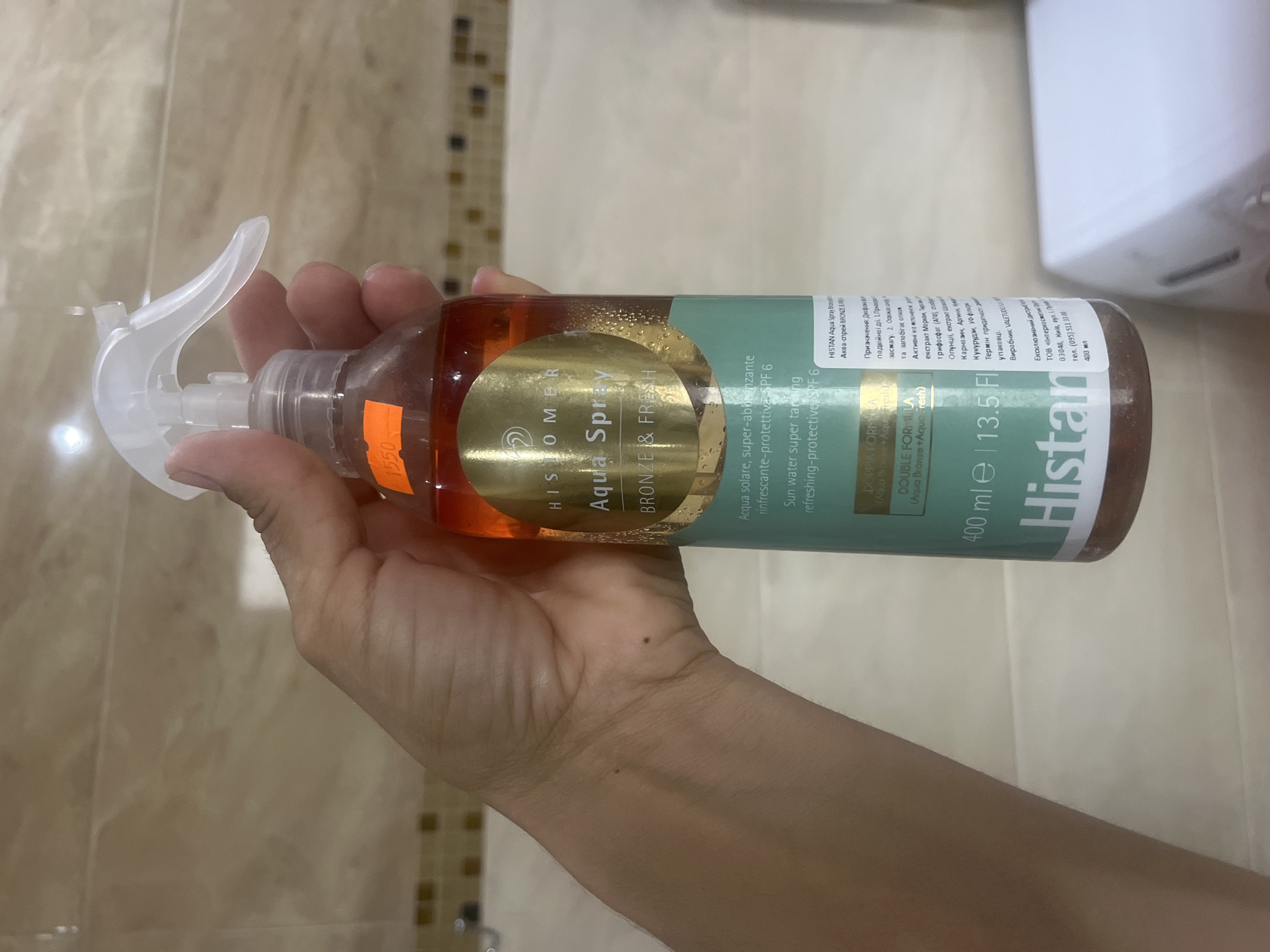 A person's hand is holding a clear plastic spray bottle filled with an amber-colored liquid. The bottle has a white spray nozzle and a white label with green and gold text. A bright orange price sticker with black numerals is affixed to the bottle. The background appears to be a tiled wall, possibly in a bathroom, with beige tiles and a mosaic border. The bottle is partially obscuring a white object in the lower right of the frame, likely an appliance such as a washing machine. The image does not contain any specific information that would allow for its localization in Onokivtsi, Ukraine.