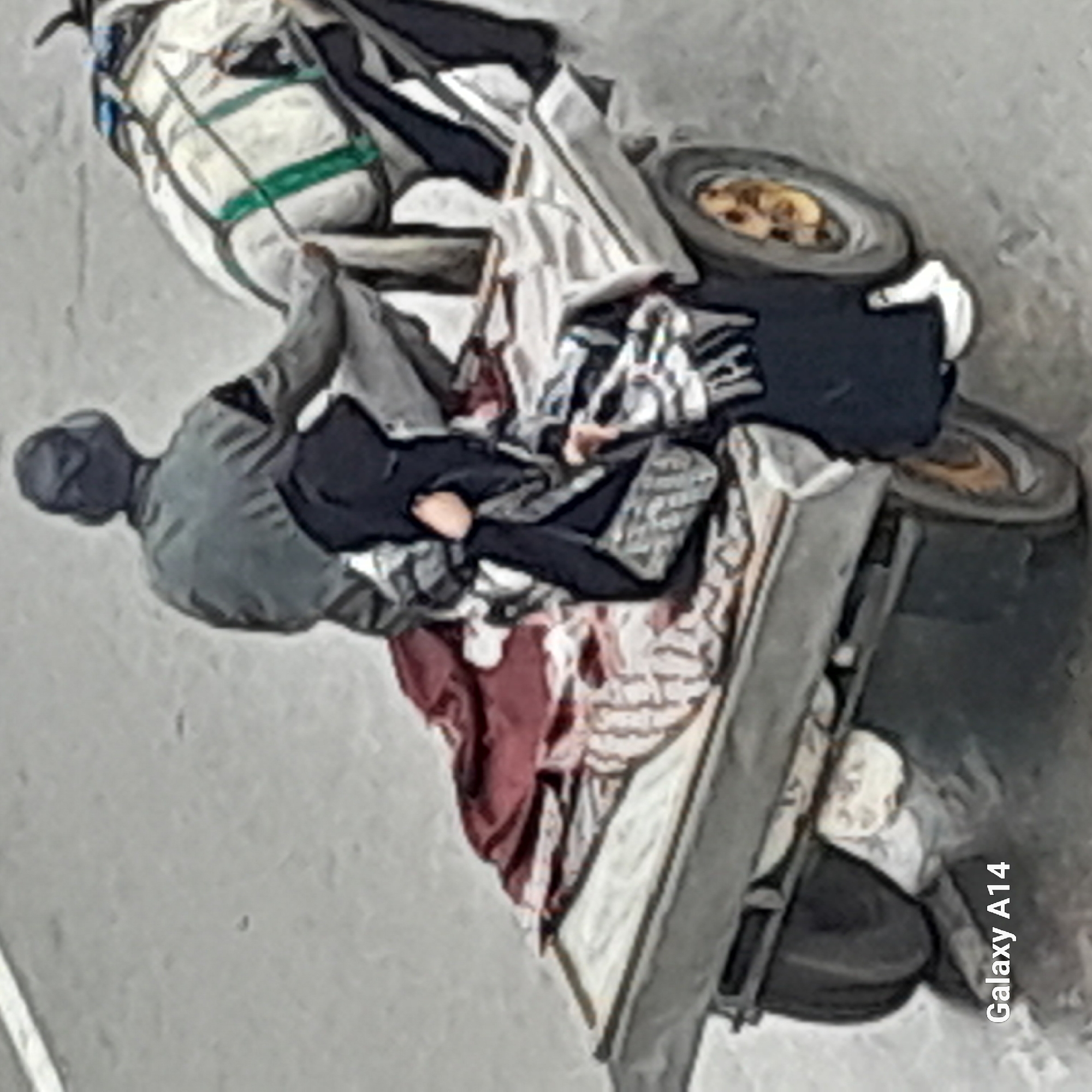 A man wearing a gray hooded jacket and dark pants pushes a two-wheeled cart loaded with belongings down a gray street. The cart is filled with assorted items including what appears to be a large white bag with green straps, various pieces of clothing in white, black, and other colors, and a burgundy-colored blanket. The wheels of the cart are visible, indicating it is in motion. The location is Unknown, Unknown. The environment consists of a paved surface.