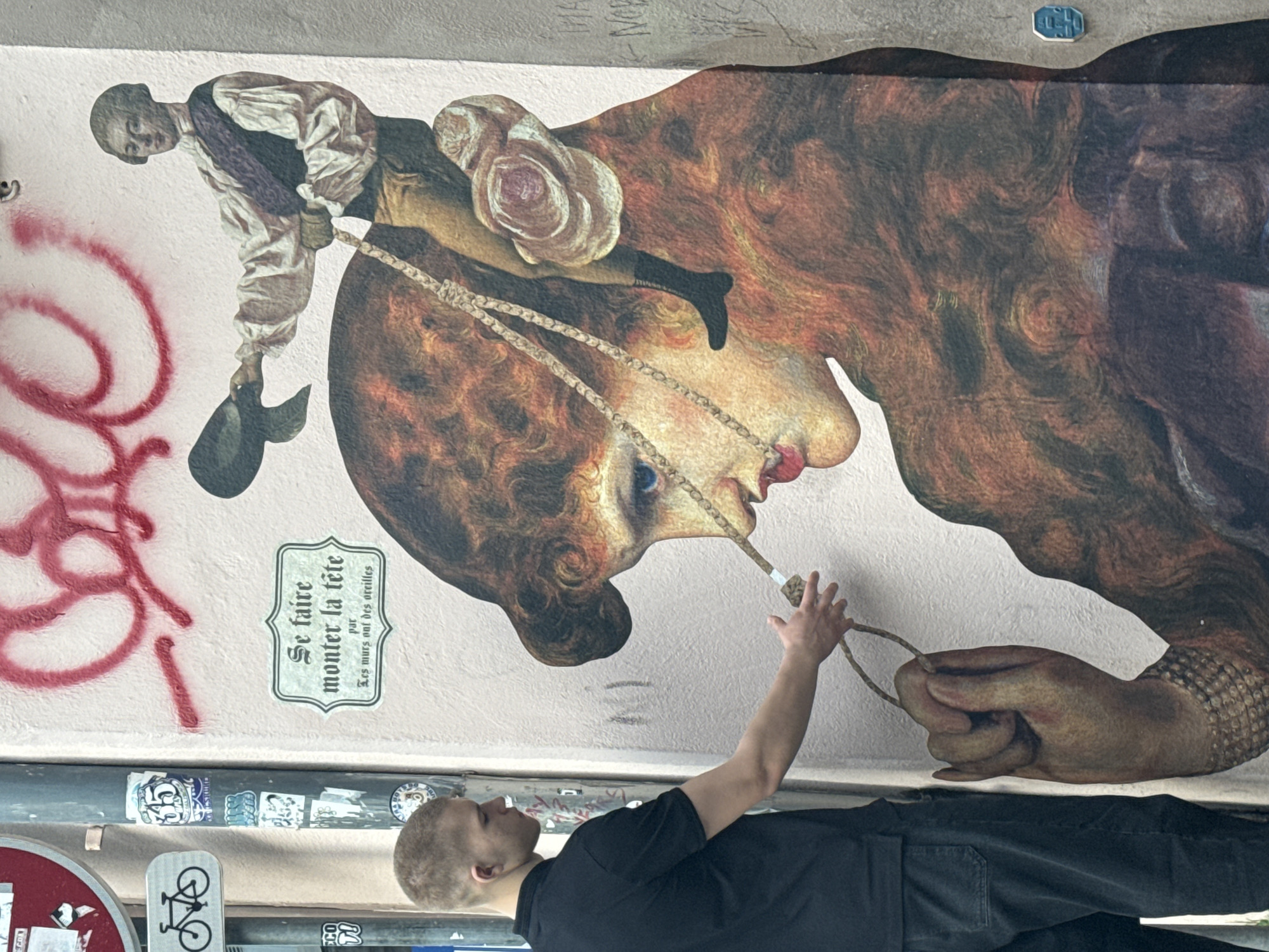 A mural in Strasbourg, France, depicts a composite image of figures. A man in modern clothing stands facing the mural. He is wearing a black shirt and dark trousers, with short, cropped hair. His right arm is raised toward the mural, his hand almost touching a rope-like detail extending from the mouth of one of the figures in the mural.
The mural features an oversized profile of a head with wavy hair, rendered in brown and orange tones. Another smaller figure is perched on top of this head, dressed in antiquated clothing. A rosette-like ornament is positioned behind their back.
Graffiti and stickers are present on the wall below the mural. A green plaque on the wall reads "Se faire monter la tête par les murs out des oreilles." Red graffiti is visible to the left. A bicycle symbol is on the bottom left.