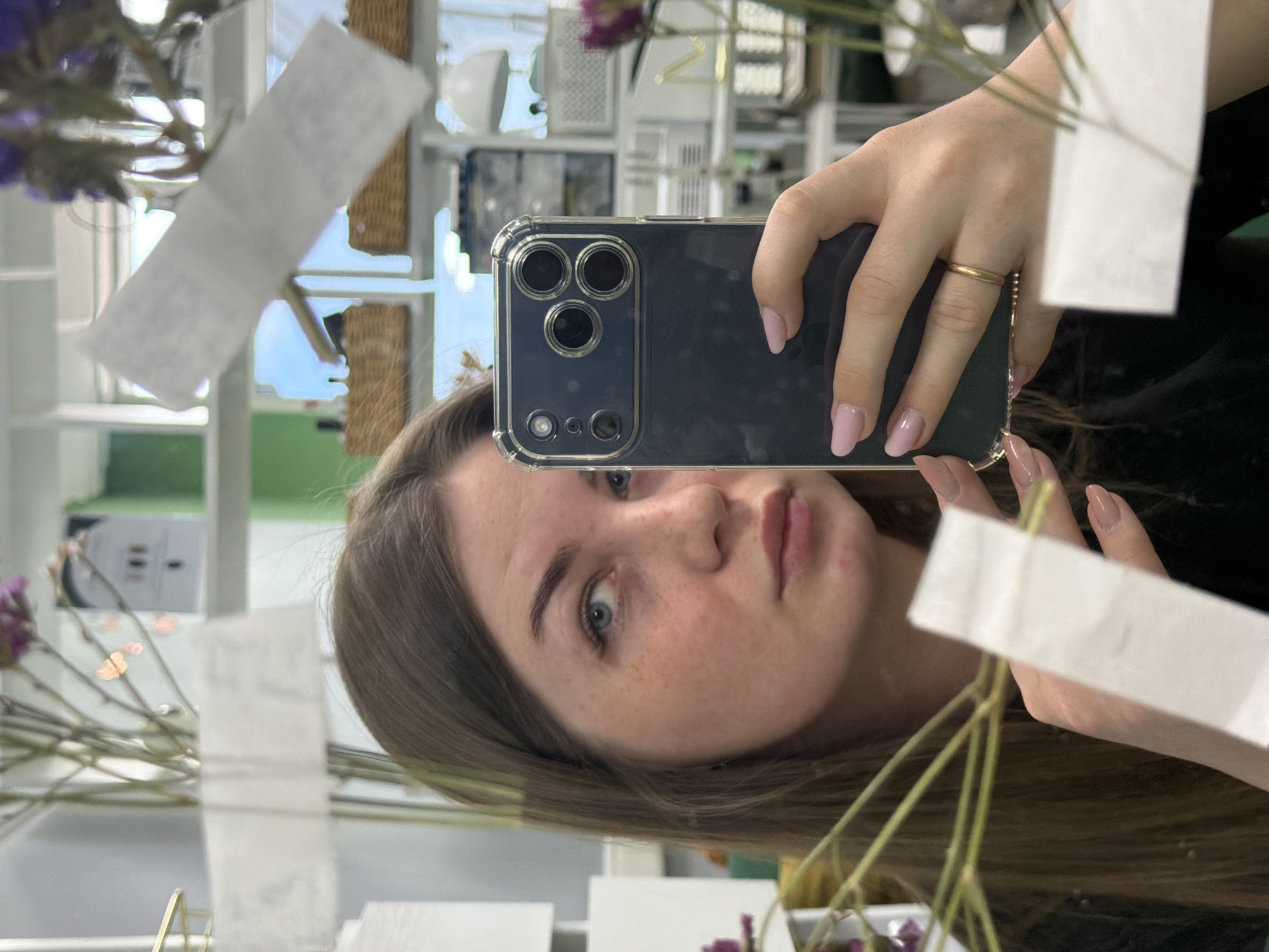 A selfie captured in a mirror reveals a young woman holding a smartphone. The phone case is transparent with a dark blue back and a triple-lens camera. The woman has light brown hair, fair skin with freckles, and light-colored eyes. She is wearing a gold ring on her finger, and her nails are painted a light pink. The background shows a room with shelves containing various items, including baskets and decorative pieces. There are also dried flowers with white tags attached to their stems in the foreground. The location is suggested to be in the city of Chaikovsky, Russia, based on inferred context not visible in the image.