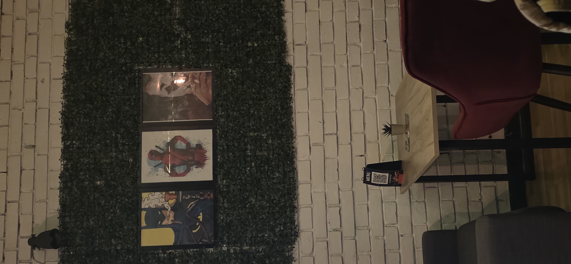 The image depicts an indoor cafe setting with a brick wall and artificial green foliage. On the wall is a triptych of framed comic book artwork. A small table with a menu and a faux succulent is situated next to a red chair and a grey sofa. The location is potentially in Sofia, Bulgaria.