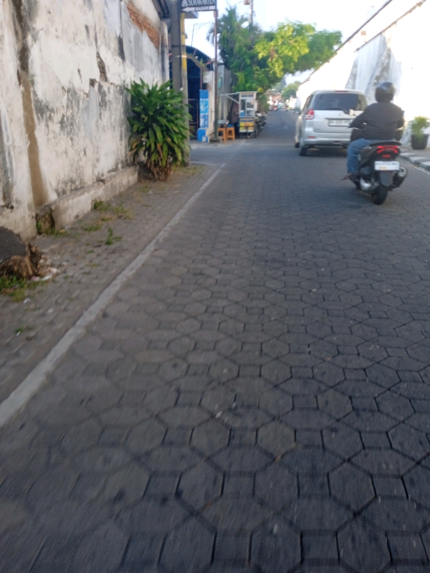 The image depicts a street scene in the city of Surakarta, Indonesia. A person is riding a dark-colored scooter, wearing a dark jacket and blue pants, and a helmet. The scooter is in motion on a paved road with a hexagonal pattern. In the background, a silver car is also moving. A building with a white, weathered wall and some brickwork is visible on the left side of the street. A lush green plant is growing against the wall. Further down the street, there are several small stalls or shops. Trees and a clear sky are visible in the distance, suggesting daylight. The overall impression is a typical urban street environment in Surakarta.