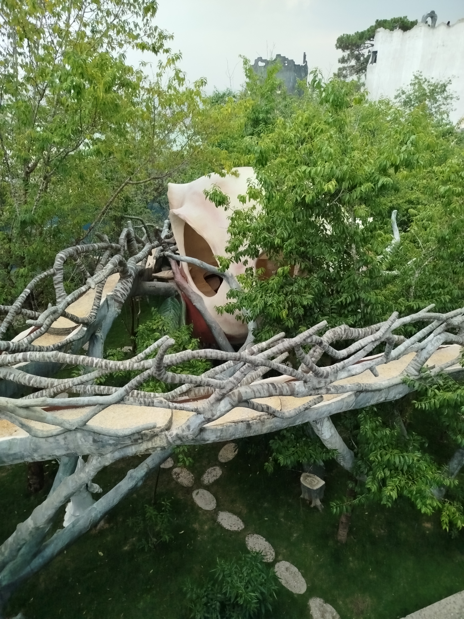The image depicts a high-angle view within Crazy House, an unconventional structure located in Da Lat, Vietnam. A winding, elevated pathway resembling tree branches dominates the foreground, constructed from grey-toned concrete. The walkway is lined with a light-colored, sandy-textured surface. Below the pathway, a grassy area is visible, interspersed with a stepping-stone path and tree stumps.
Behind the walkway is a large, organic-shaped structure, partially obscured by foliage. This structure is painted in muted flesh tones, with prominent openings suggesting it is intended to resemble an animal form.
Lush green trees and foliage fill the space surrounding the structures, creating a dense, natural environment. In the distance, glimpses of other buildings can be seen, including a dilapidated structure and a white building with greenery on top. The sky is overcast, providing soft, diffused lighting throughout the scene. There are no visible people or interactions in the image.