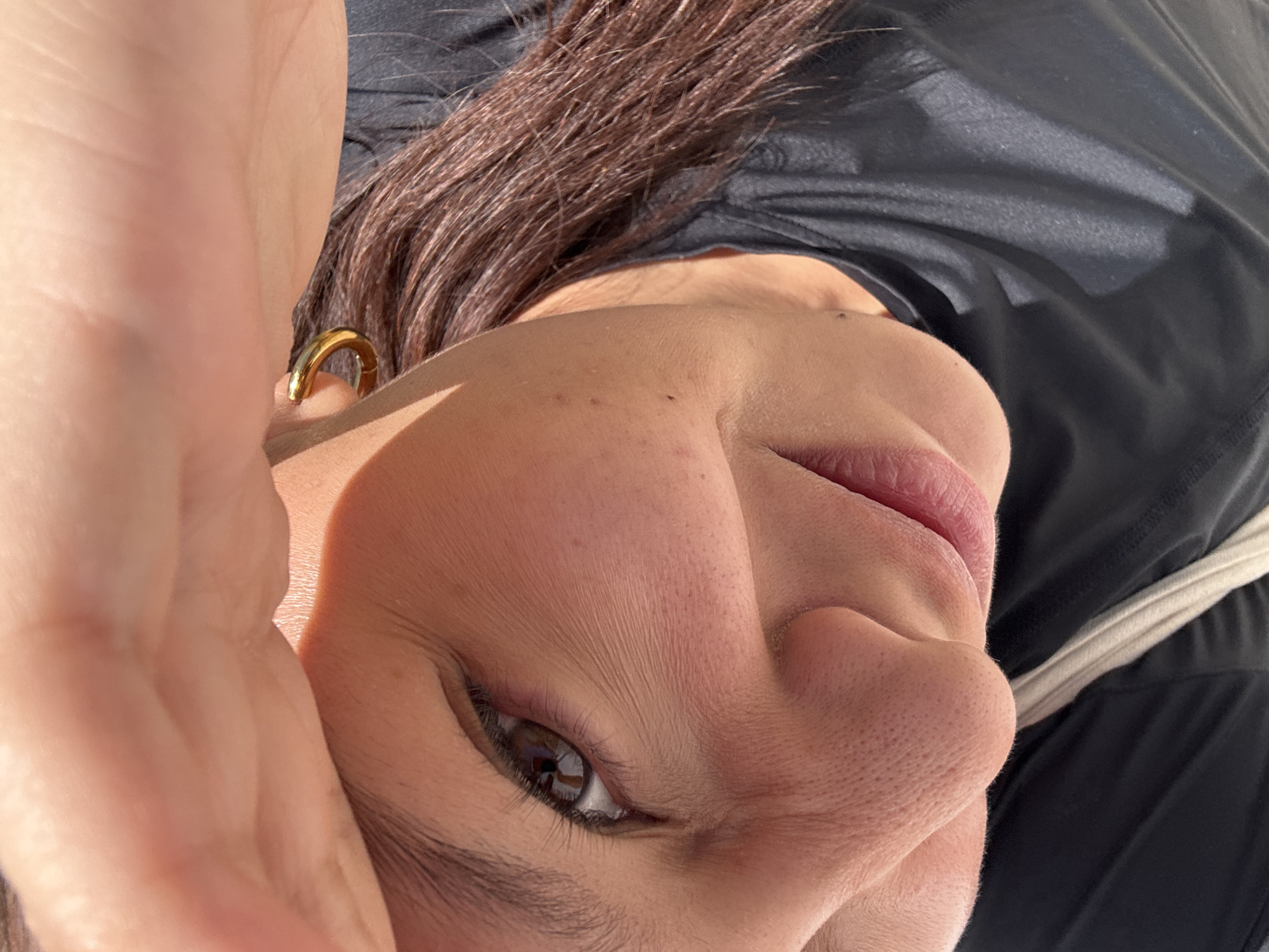 A close-up, high-angle portrait captures the partially obscured face of a person, likely a woman, in San Andrés Cholula, Mexico. The subject's head is tilted, with her gaze directed upwards and towards the camera, suggesting a selfie or a relaxed pose. Her left hand is visible in the foreground, partially covering the top left of the frame, with strong sunlight highlighting its contours.
The person has brown eyes, visible through thick eyelashes, with a reflection of a window or light source evident in the iris. Her lips are slightly parted, revealing a natural pink hue, and her nose is prominently featured due to the angle. Her skin shows natural texture, including visible pores, and appears to be illuminated by direct, bright sunlight, creating strong highlights and shadows across her face.
Long, dark brown hair flows from the top right towards the bottom left of the frame, partially covering her shoulder and neck area. A gold hoop earring is visible in her left ear, catching the sunlight. She is wearing what appears to be a black top or t-shirt, characterized by its dark, somewhat shiny fabric. A white strap, possibly from a bag or another garment, can be seen near her right shoulder. The bright lighting suggests it is daytime with clear weather. There is no visible text in the image.