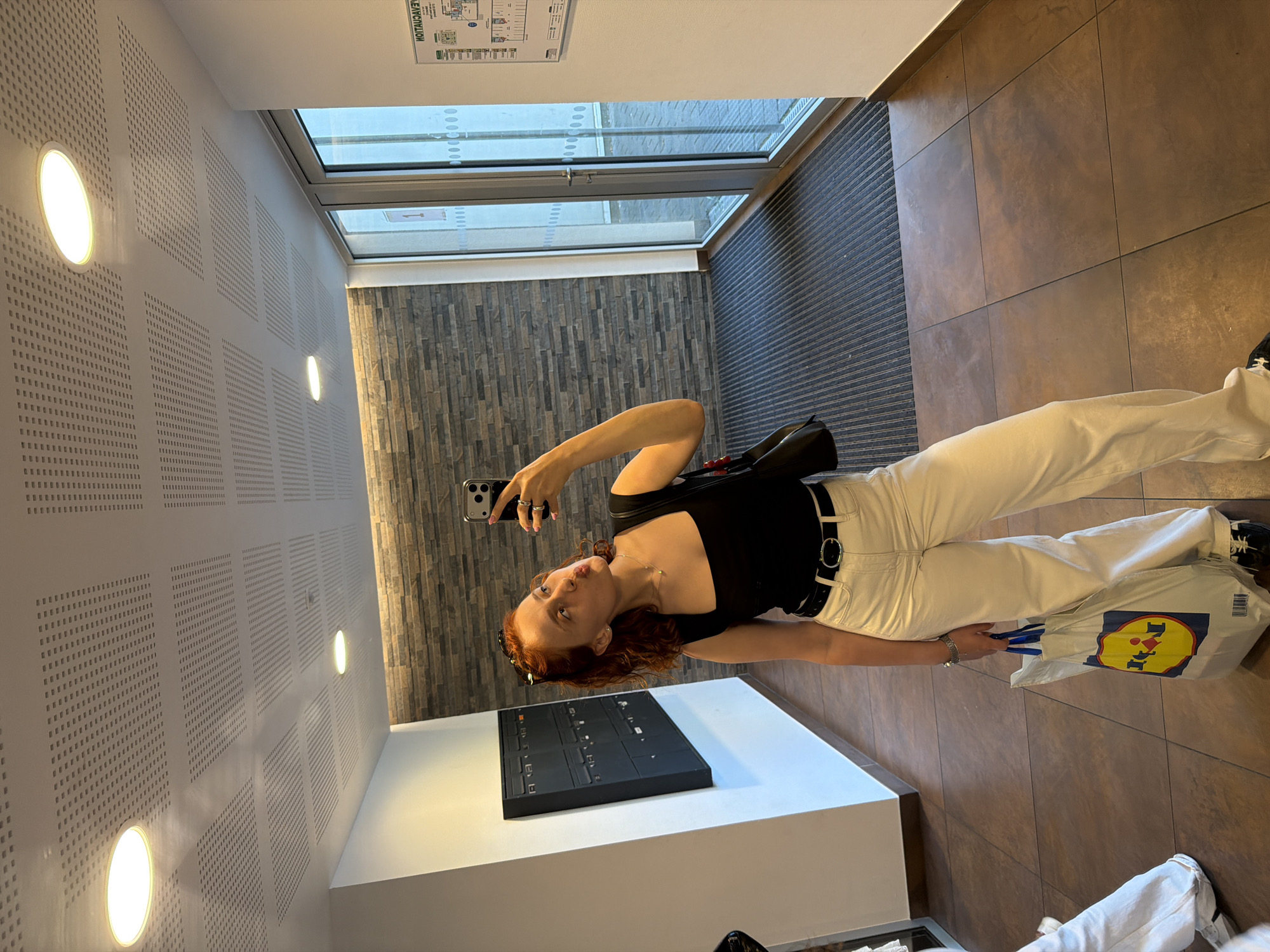In Strasbourg, France, a young woman with auburn hair is taking a selfie in a hallway. She is wearing a black tank top, white wide-leg pants, and a black belt with a silver buckle. She has a black purse slung over her shoulder and is holding a white shopping bag with the Lidl logo. The hallway has tiled floors, a stone accent wall, and a drop ceiling with recessed lighting. To the left, a black mailbox unit is mounted on the wall. To the right, glass doors lead outside. A woman in the background is partially visible, wearing white pants.