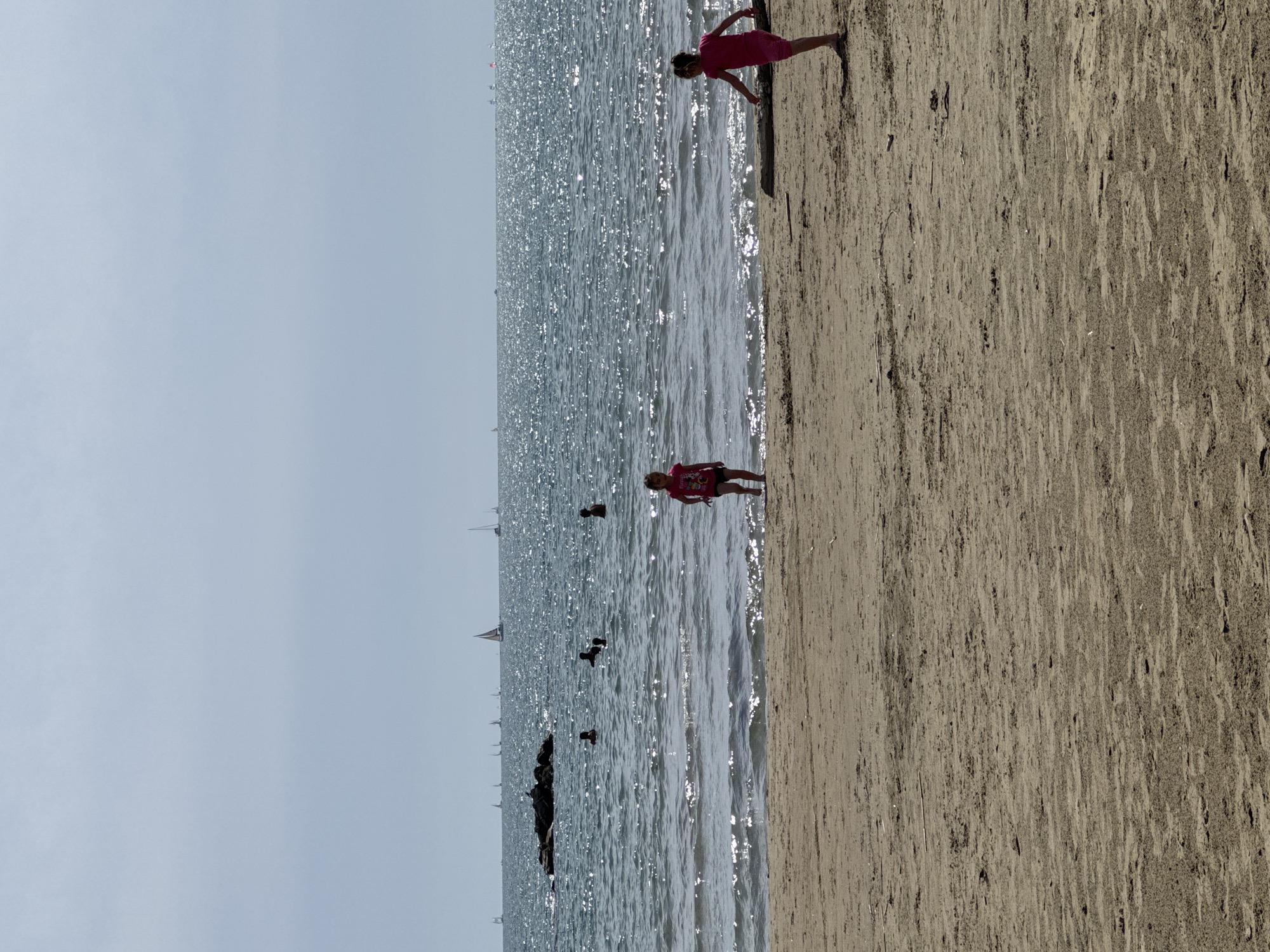 The image shows a beach scene in Nettuno, Italy. The beach consists of light brown sand and is adjacent to a body of water that reflects sunlight. Several people are visible: One child stands on the edge of the water, and another child stands on the dry sand. Some people are swimming in the water. Sailboats are in the background. The sky is overcast.